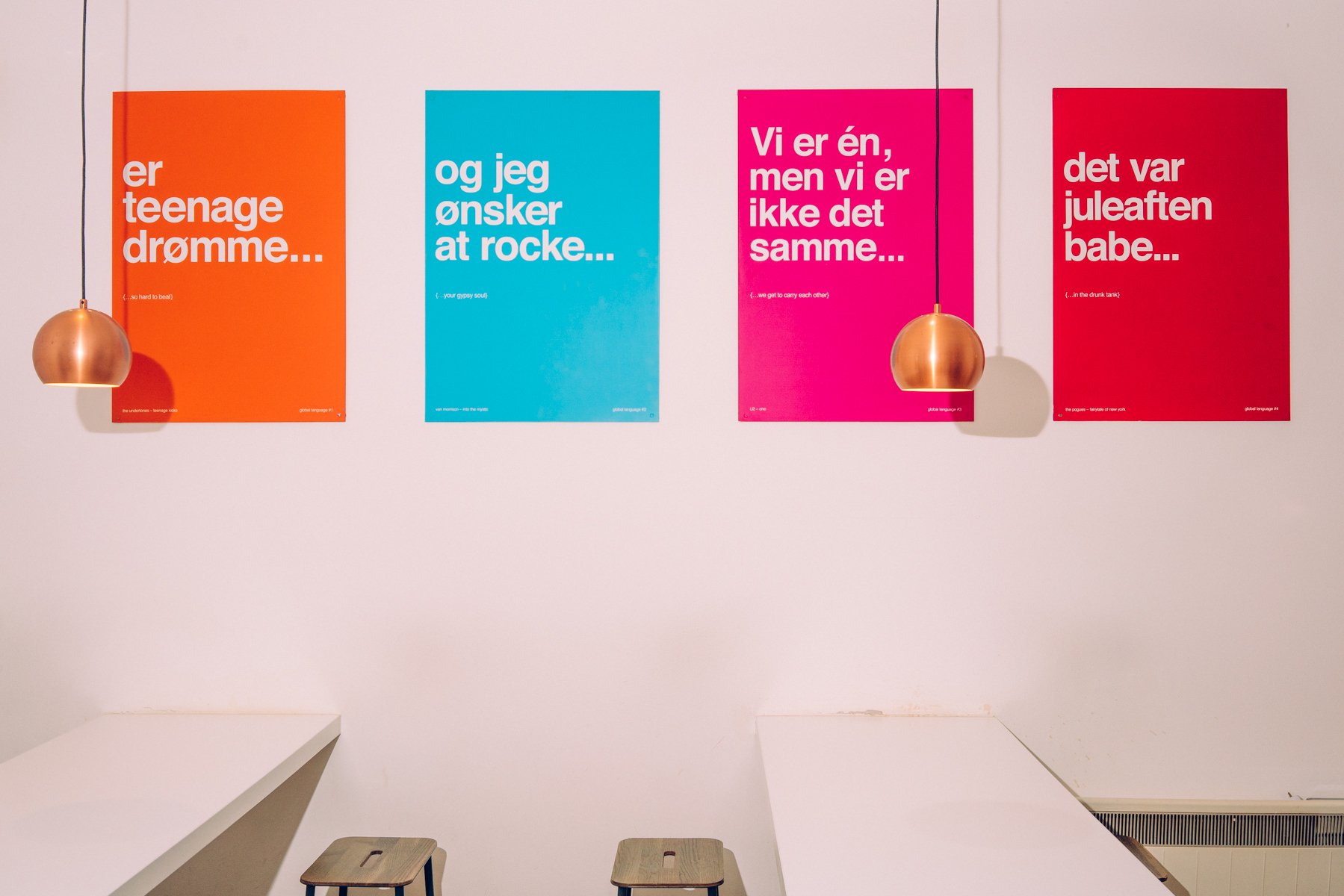 Four colorful posters with text in Norwegian and English, hanging on a white wall, illuminated by two pendant lights, with white tables and wooden stools below.