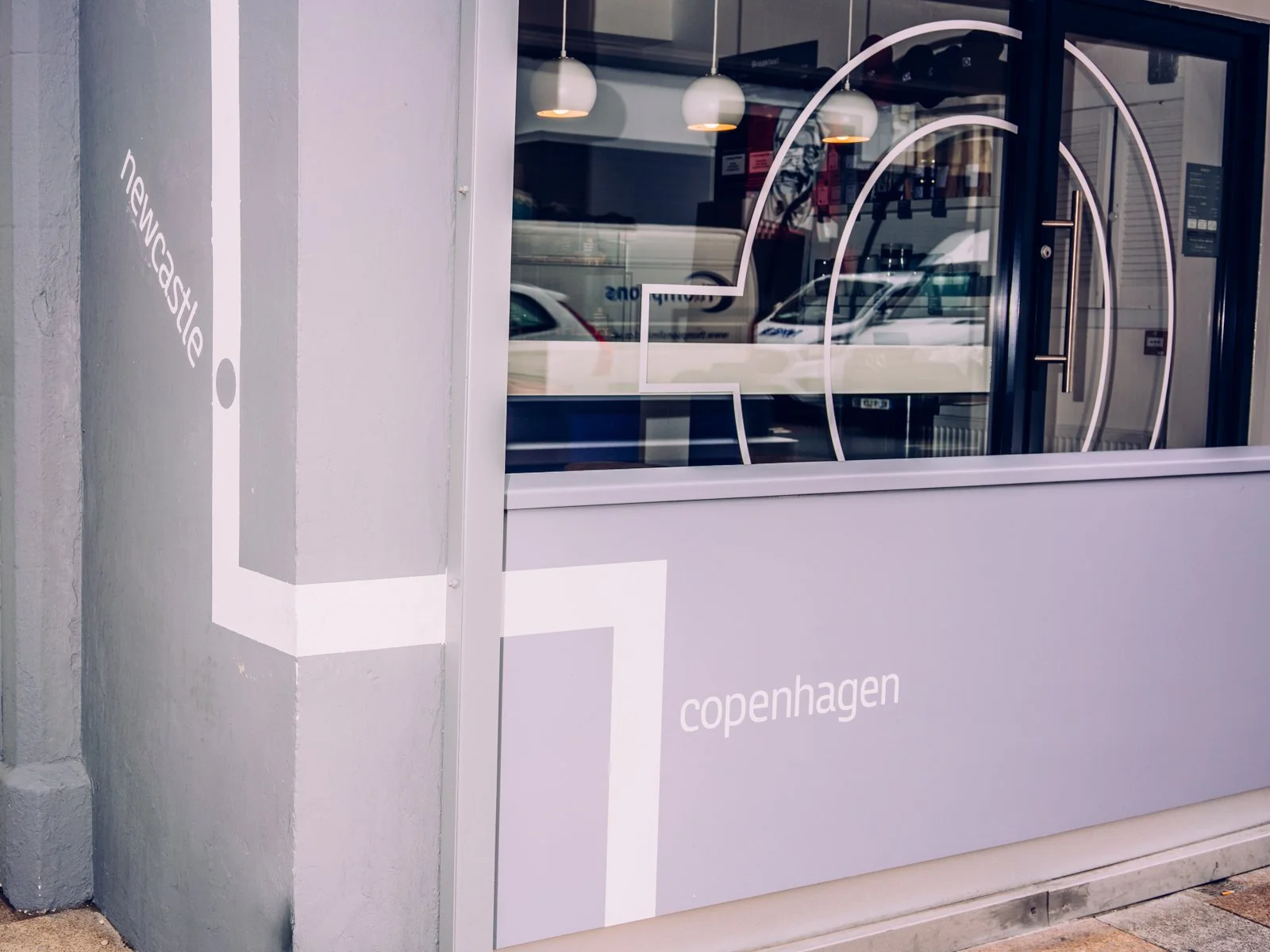 Storefront window with large white cafe logo and the word 'Copenhagen' written on the lower part of the window. Reflections of cars and street scene can be seen in the glass.