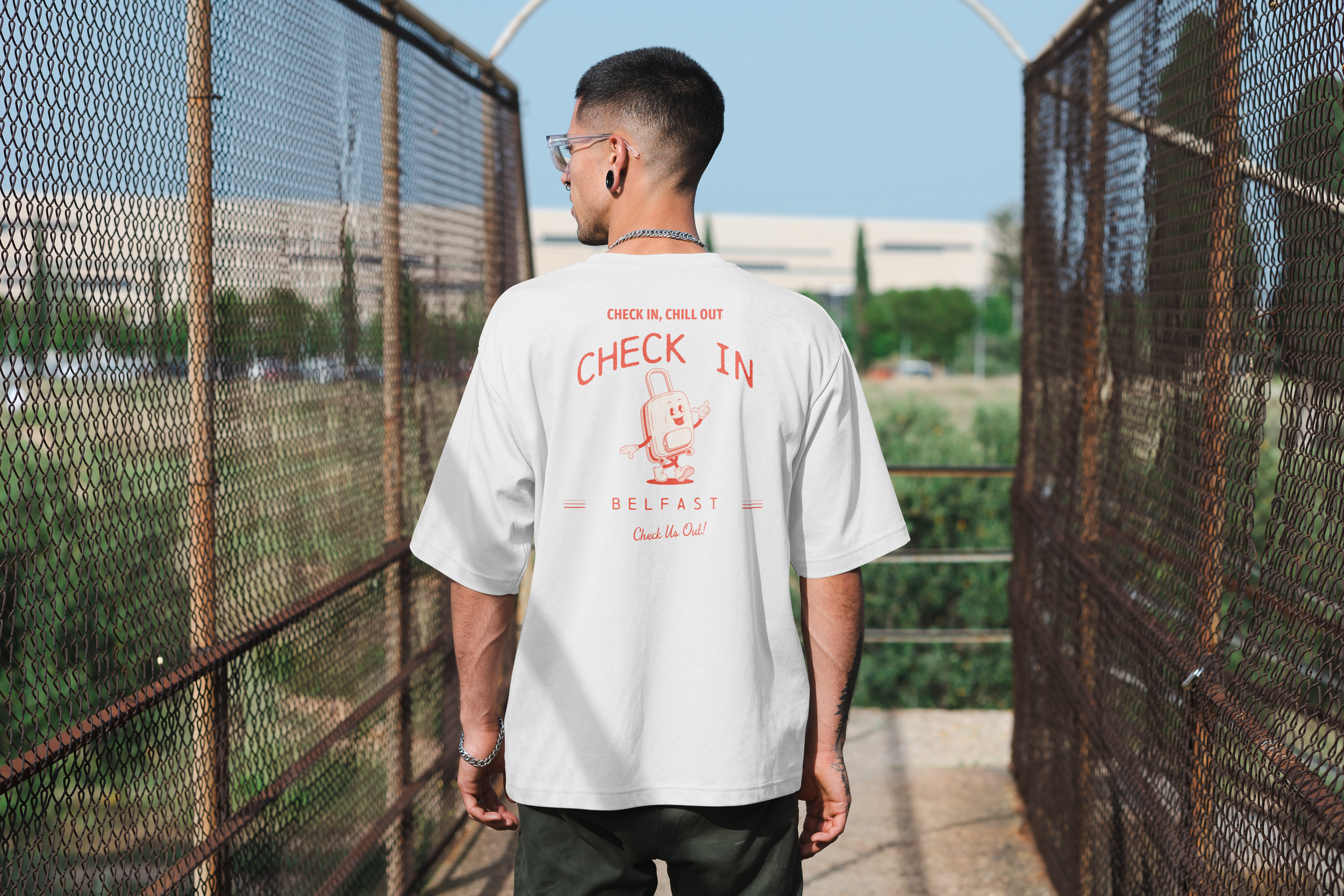 A person with short dark hair, wearing glasses, earrings, a silver chain, and a white T-shirt with a graphic of a smiling backpack and the words "Check In, Chill Out" and "Belfast" on the back, walking between chain-link fences on a sunny day.