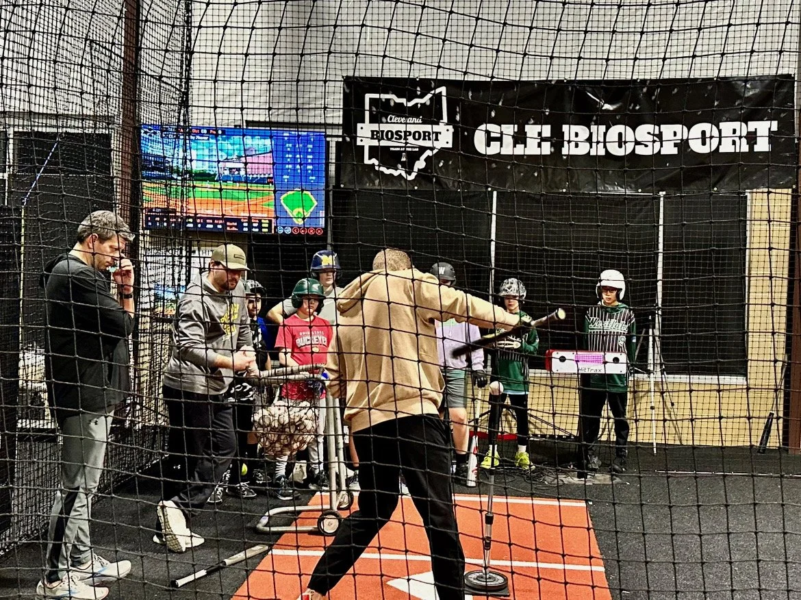 Group of boys in a baseball training facility with bats, wearing jerseys and sports gear, standing in front of a Biosport banner.