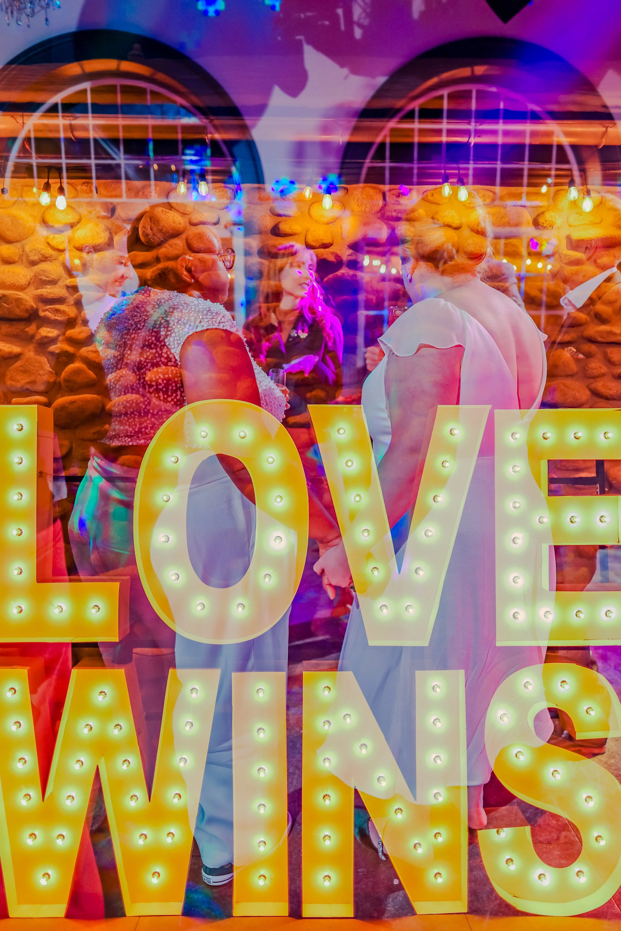Colorful illuminated 'LOVE' sign with a reflection of a group of women talking inside a venue with stone walls and bunting flags hanging.