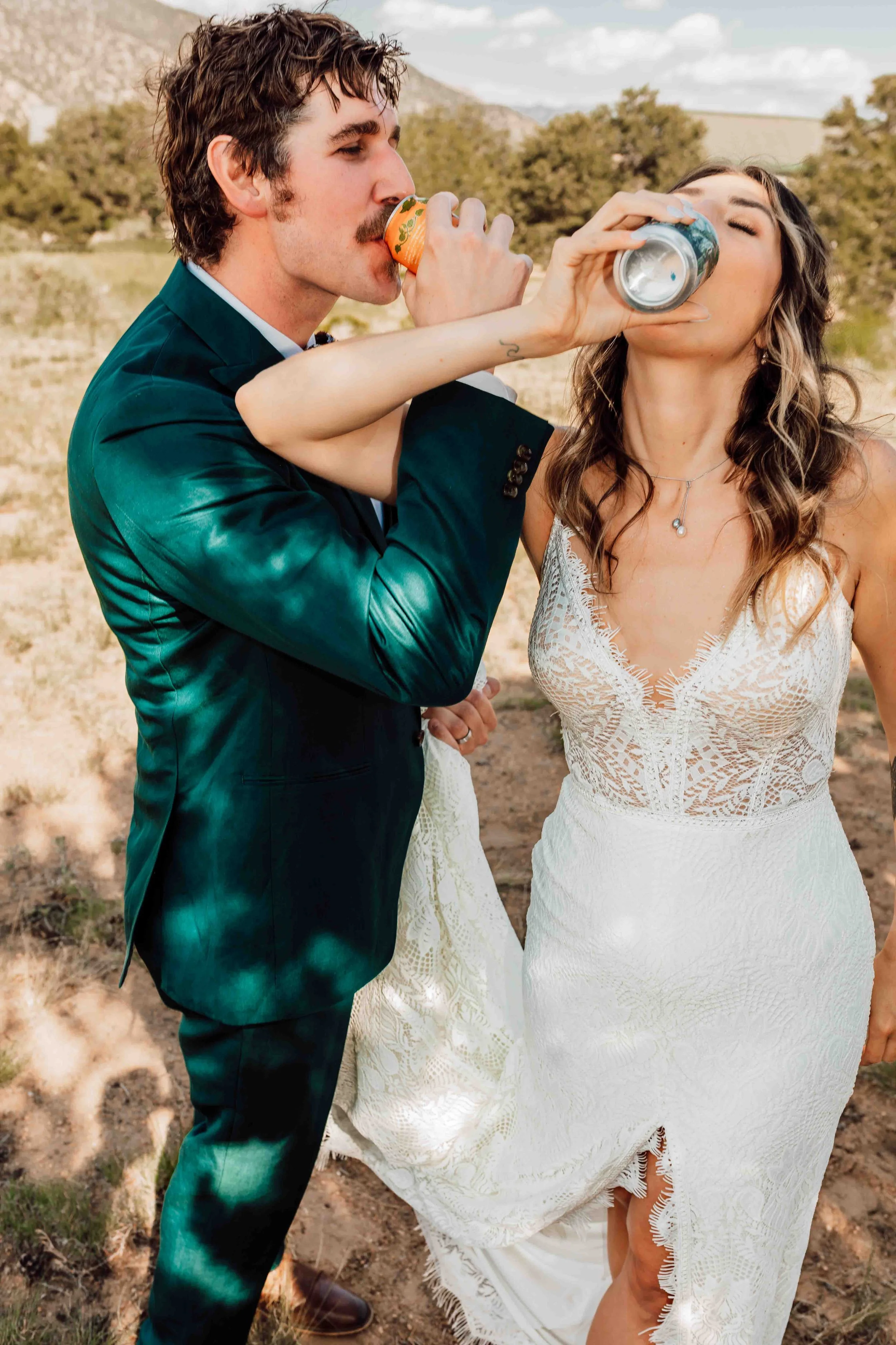 A man in a dark suit and a woman in a white lace dress are outdoors, holding cans and drinking together at sunset or daytime, with a landscape of trees and hills in the background.