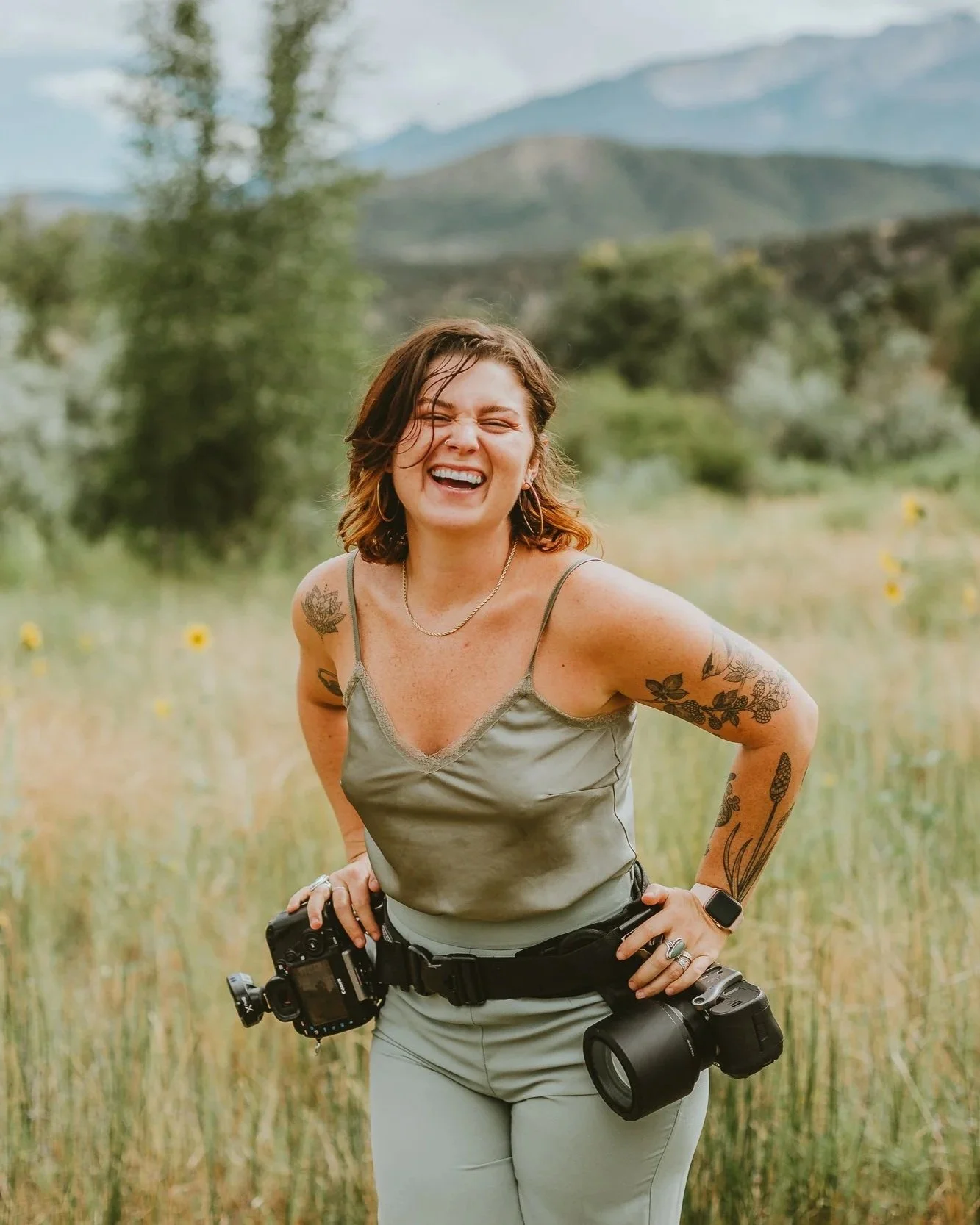 A woman with short brown hair, tattoos on her arms, wearing a gray camisole and light gray pants, smiling and posing outdoors in a grassy field with mountains and trees in the background.