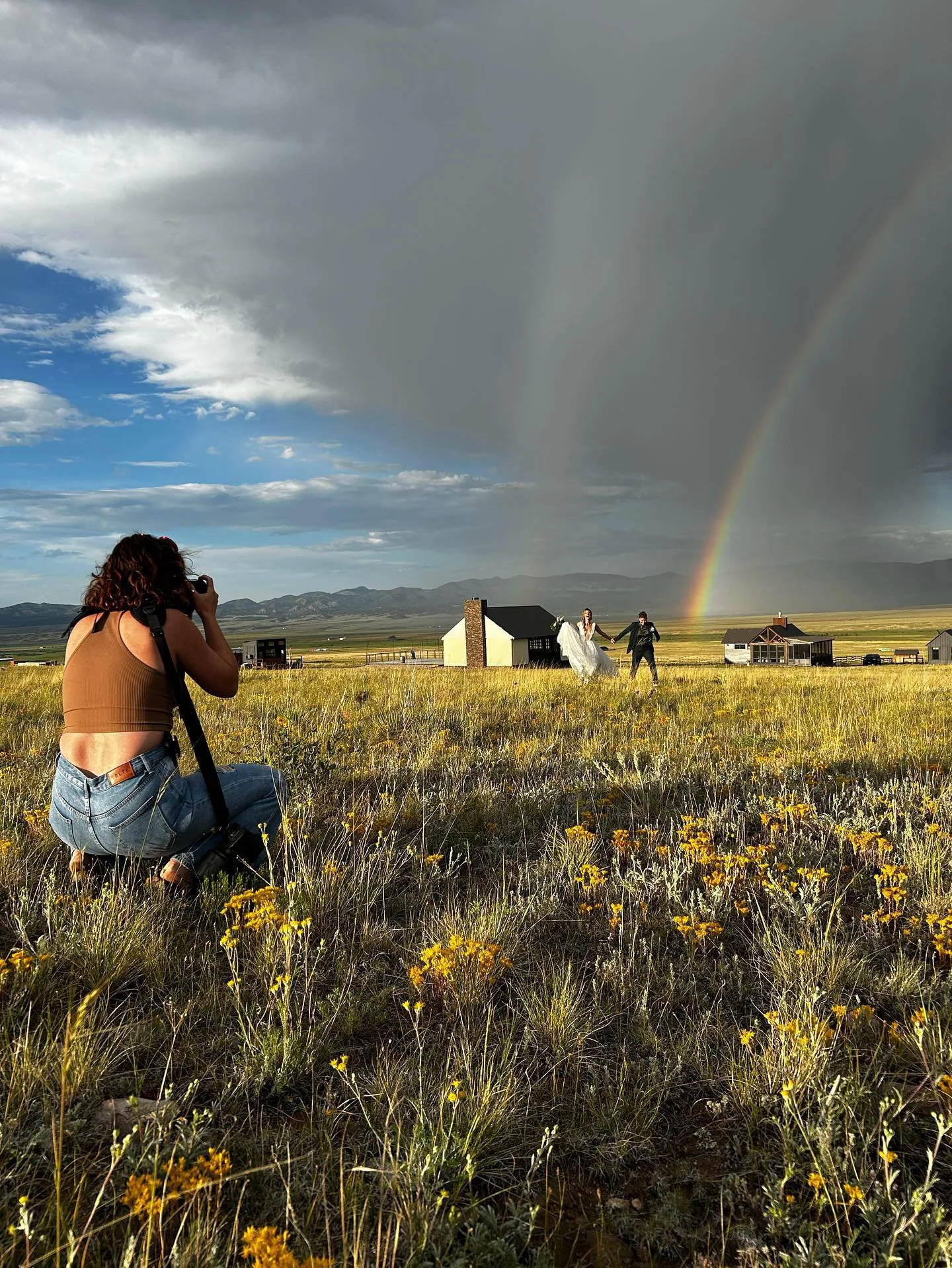 A woman with curly hair wearing a tan crop top and jeans is kneeling in a field of yellow flowers, taking a photo of a couple holding hands and walking towards a rainbow in a cloudy sky, with mountains in the background.