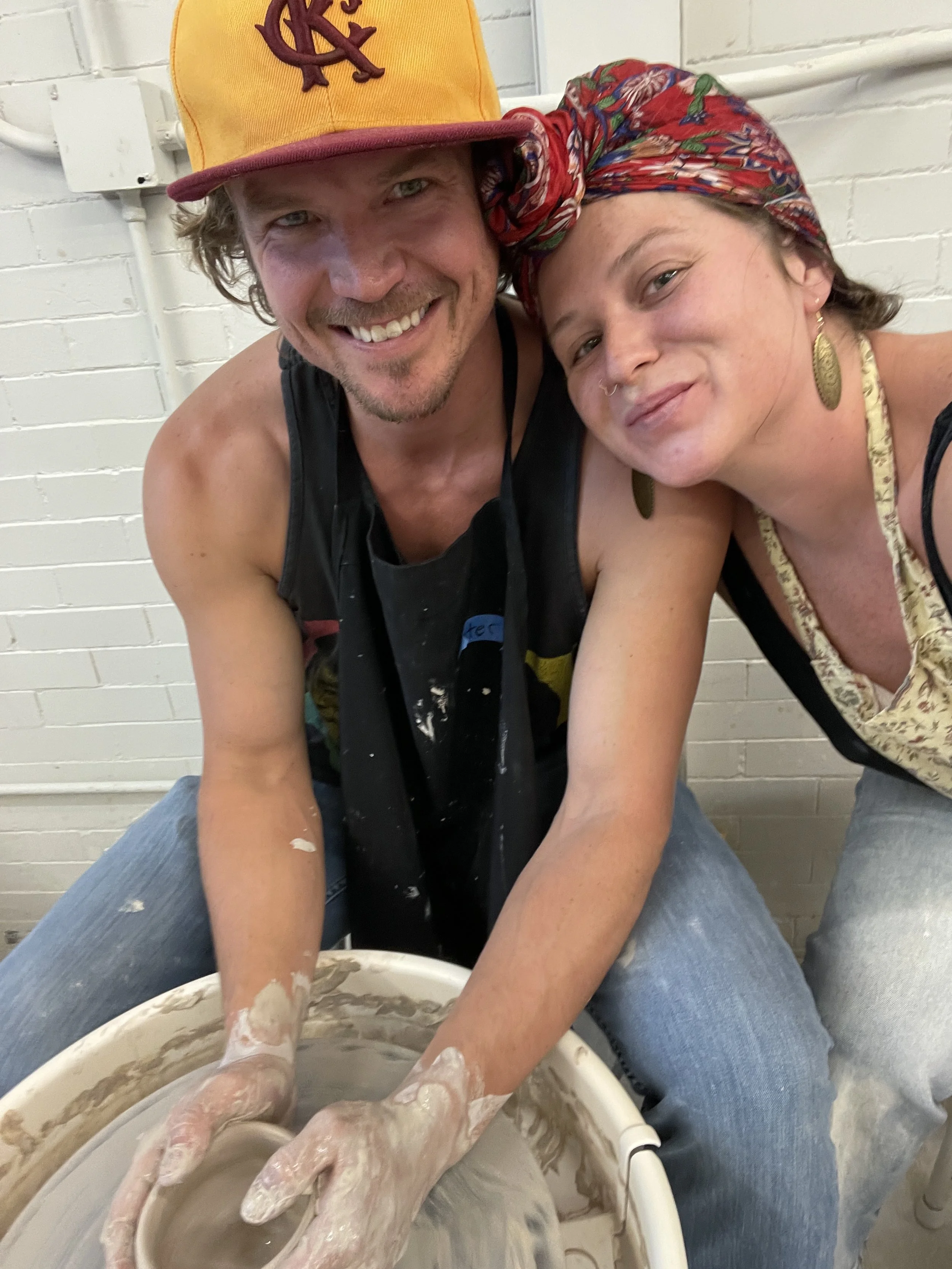 Two people working together on a pottery wheel in a studio, one person with a colorful headscarf and the other wearing a yellow and red cap, both with hands covered in clay.