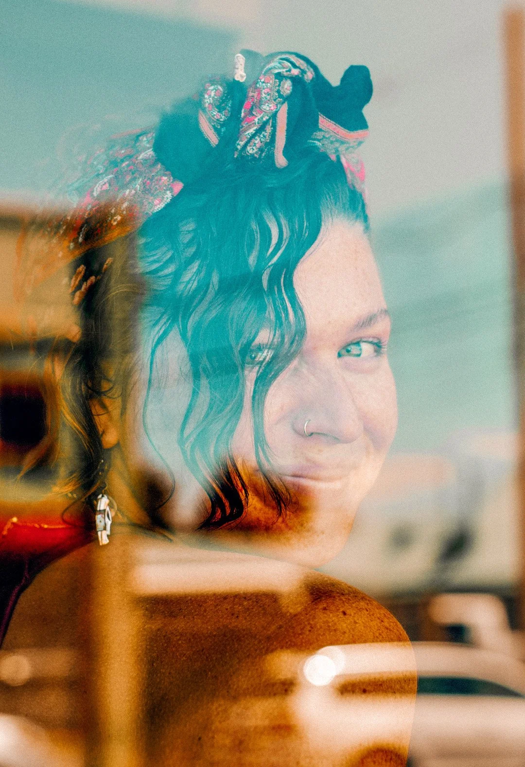 A young woman with curly dark hair, a nose ring, and earrings looks through a window, creating a layered, reflective image.