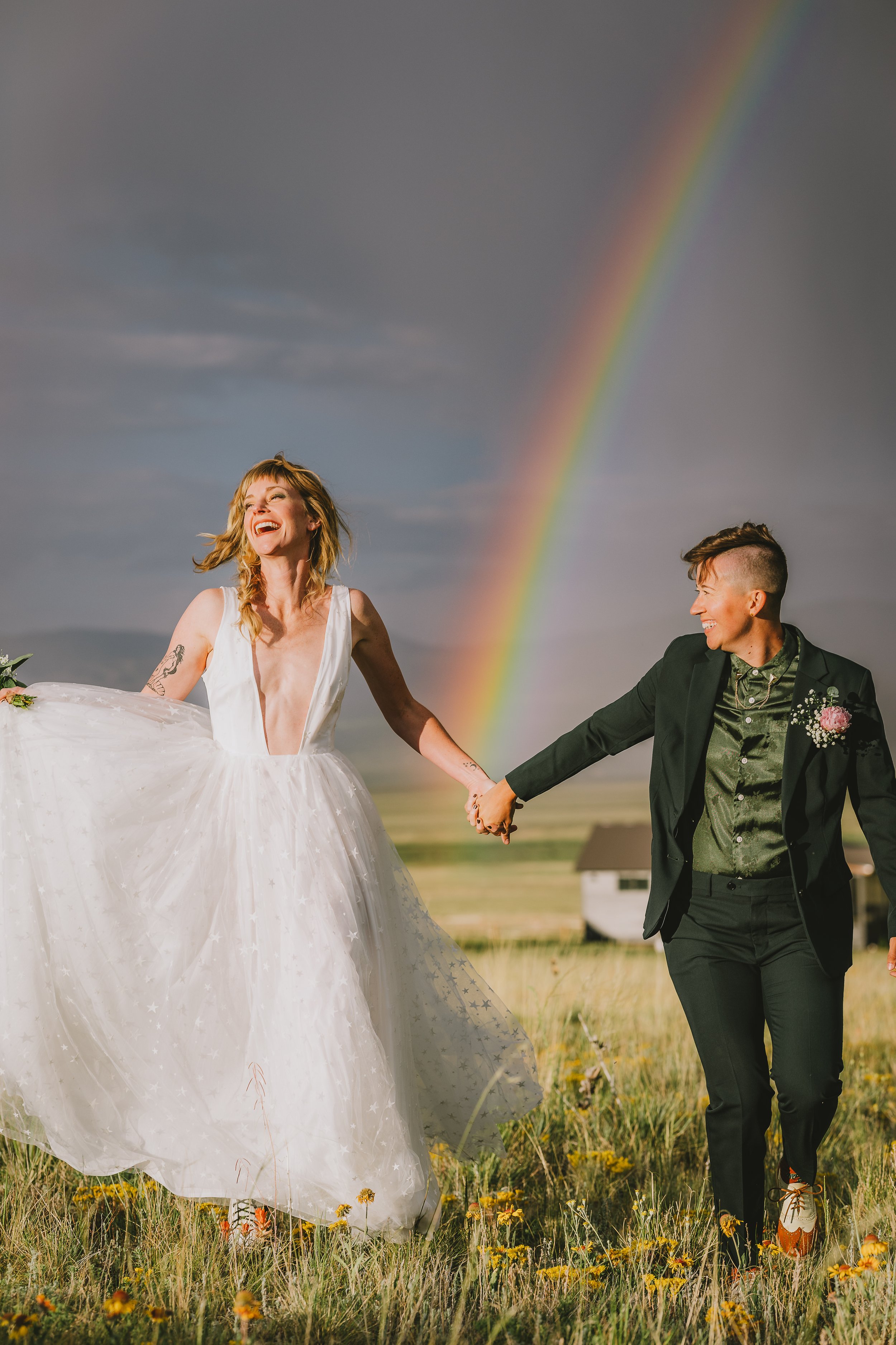 A joyful bride and groom holding hands in a grassy field with a rainbow in the background. The bride is wearing a white wedding dress, and the groom is dressed in a black suit with a green shirt. Both are smiling and looking at each other.