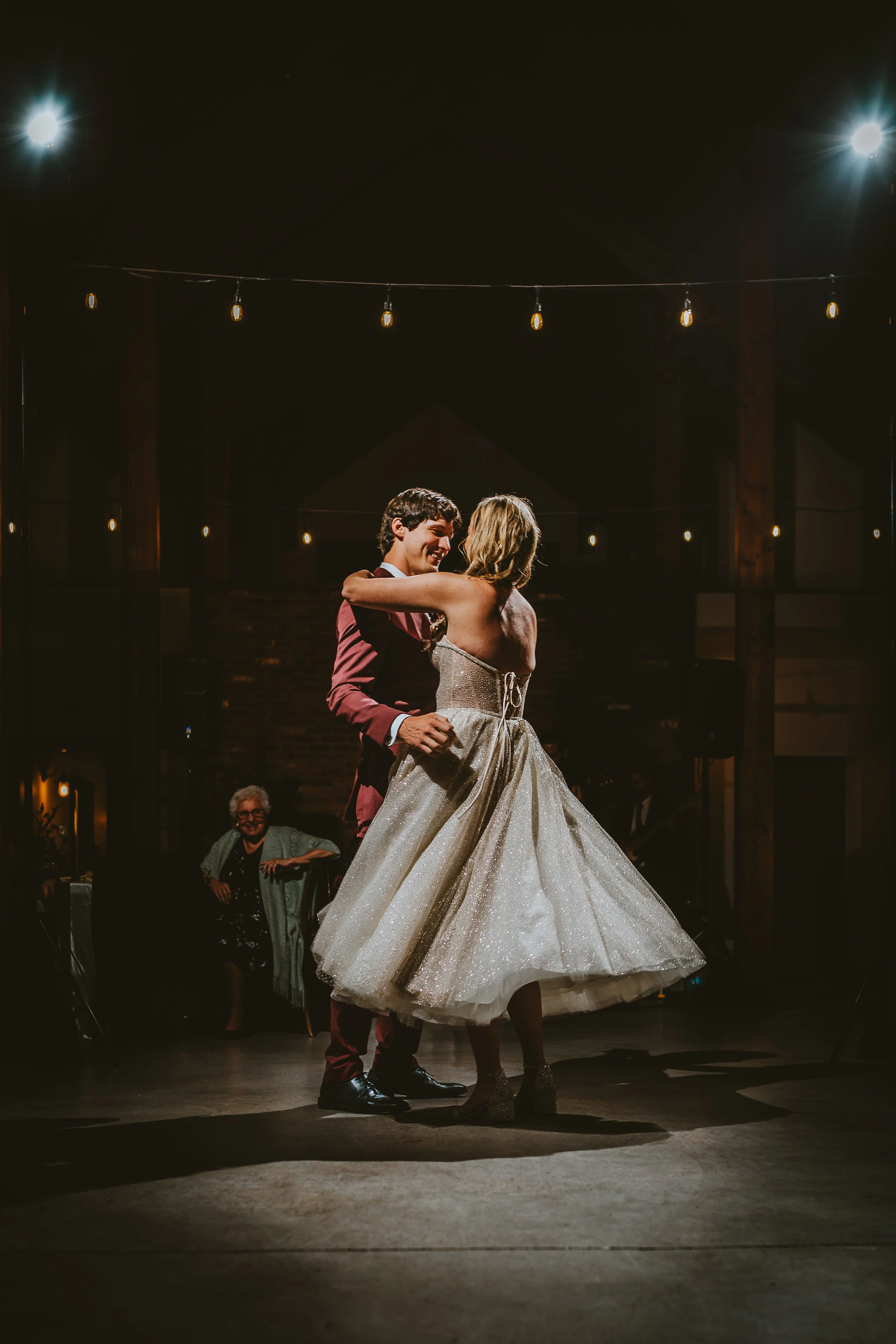 A bride and groom dancing closely during their wedding reception under string lights in a dimly lit venue. The bride is wearing a shimmering strapless gown, and the groom is in a burgundy suit with black shoes. An older woman with glasses is smiling in the background.