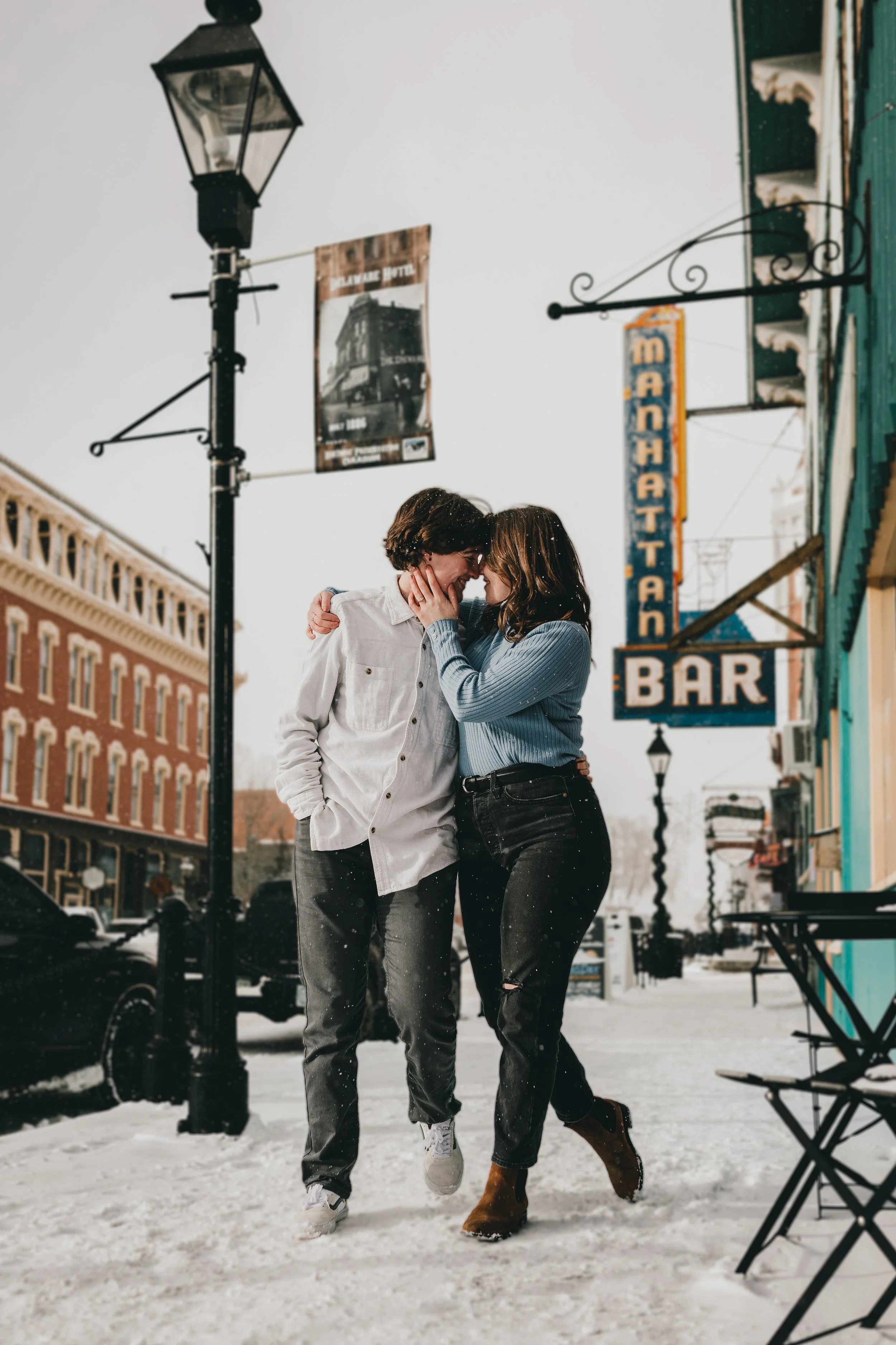 A young couple with light skin and dark hair, smiling and embracing each other, walking on a snowy city sidewalk, with vintage buildings, street lamps, and signs in the background.