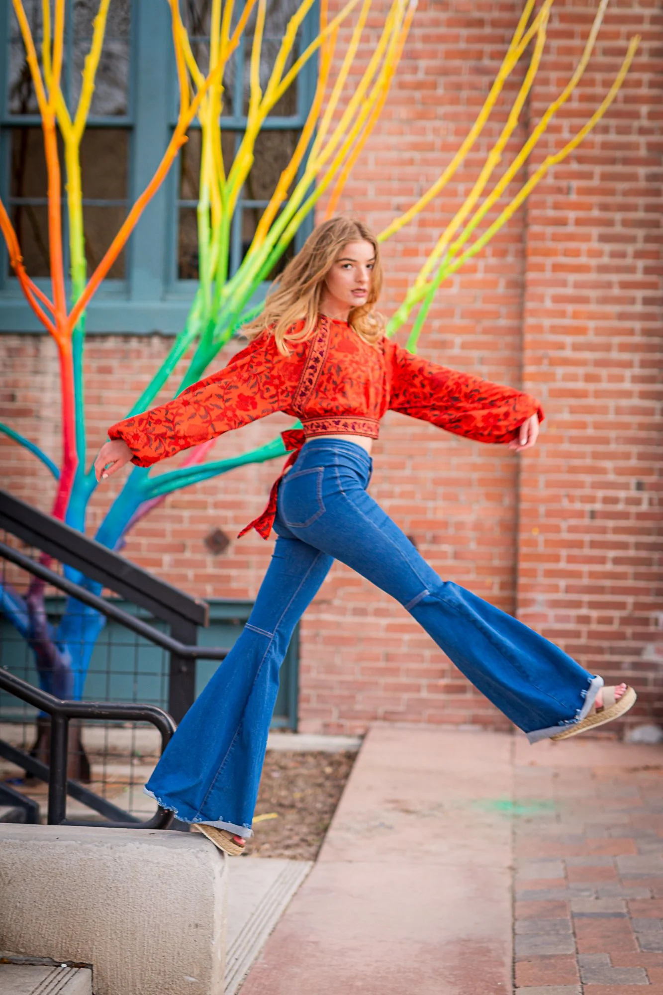A young woman is captured mid-motion on a staircase outdoors, wearing a red patterned blouse and blue flared jeans, with her arms outstretched and one leg extended. Behind her is a brick building with a window, and an abstract, colorful sculpture resembling tree branches.