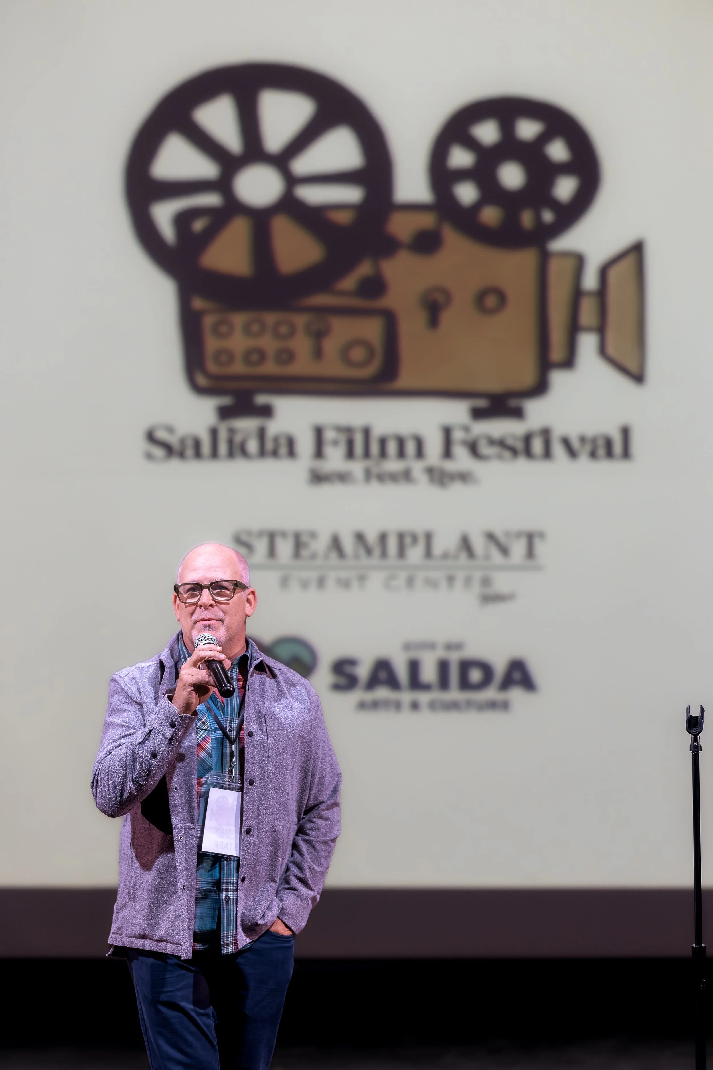 A man in glasses and a gray jacket with a checkered shirt and lanyard speaking into a microphone on stage with a large blurred poster in the background that reads 'Salida Film Festival'.