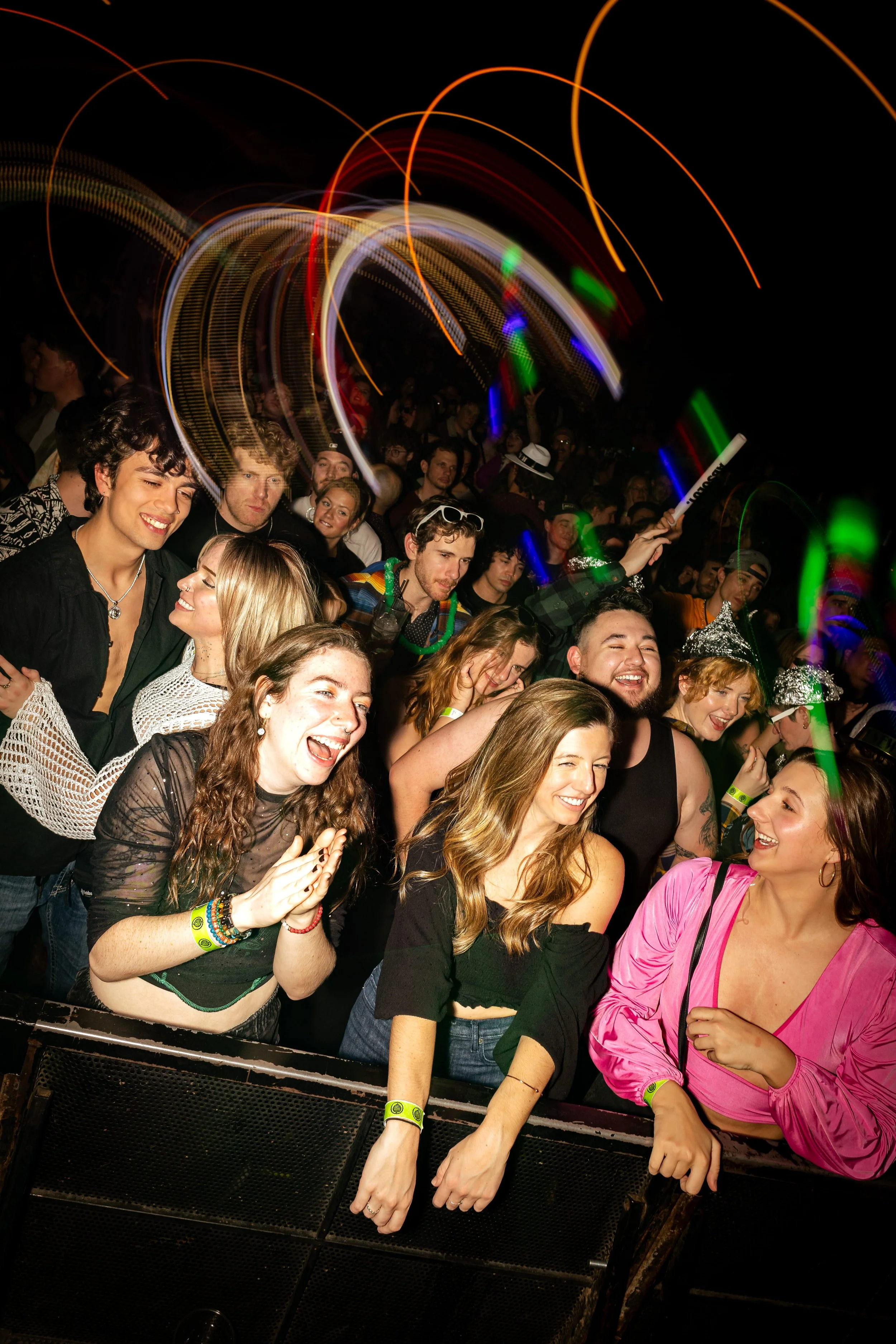A crowd of people at a concert or festival, many smiling and enjoying the event, with colorful lights and motion streaks in the background at night.