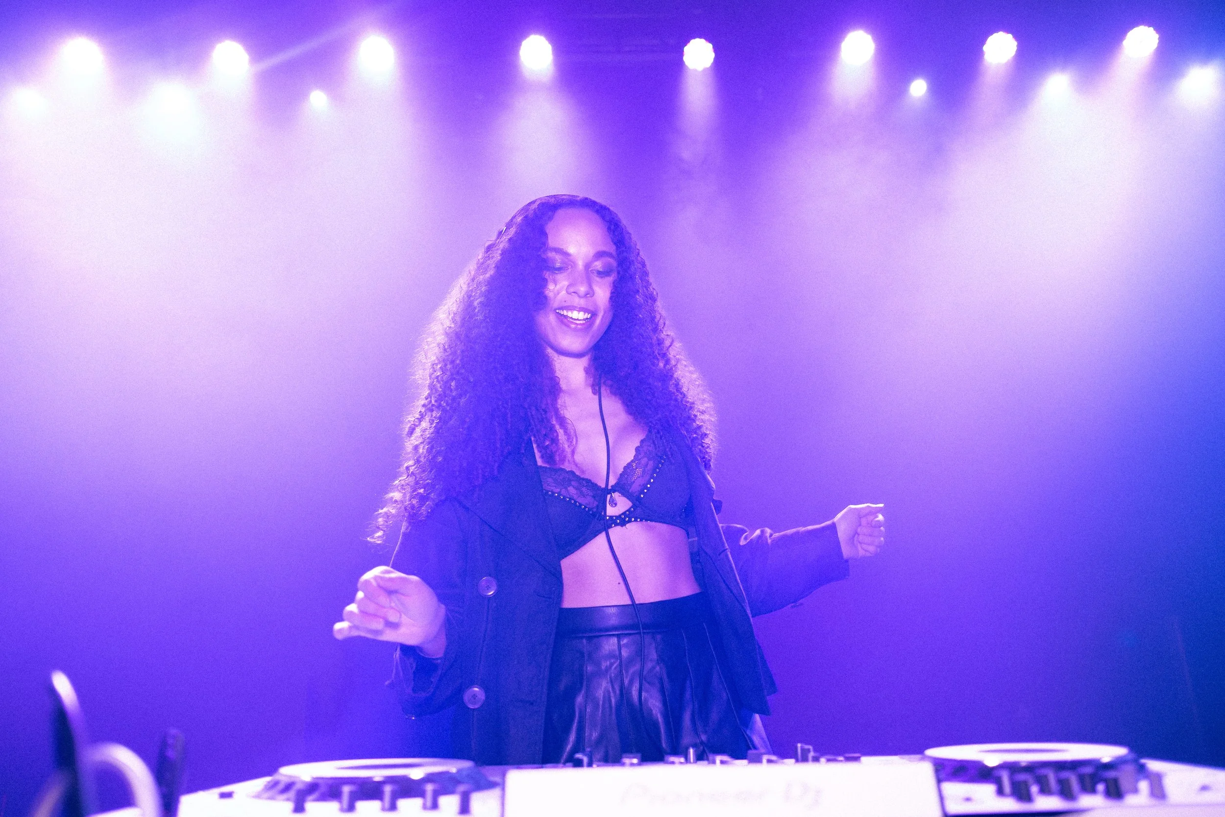 A woman DJ with curly hair wearing a black lace bralette and a black jacket, smiling and dancing under purple stage lights, with a DJ mixer in front of her.