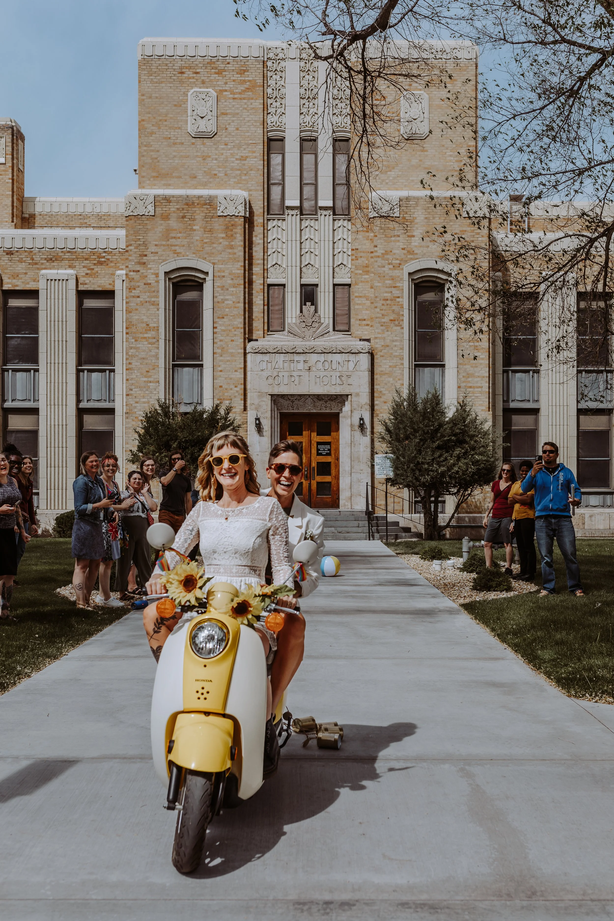 A woman riding a yellow motor scooter with sunflowers and rainbow ribbons, leading a group of smiling people in front of a historic courthouse building, with leafless trees and a clear sky in the background.