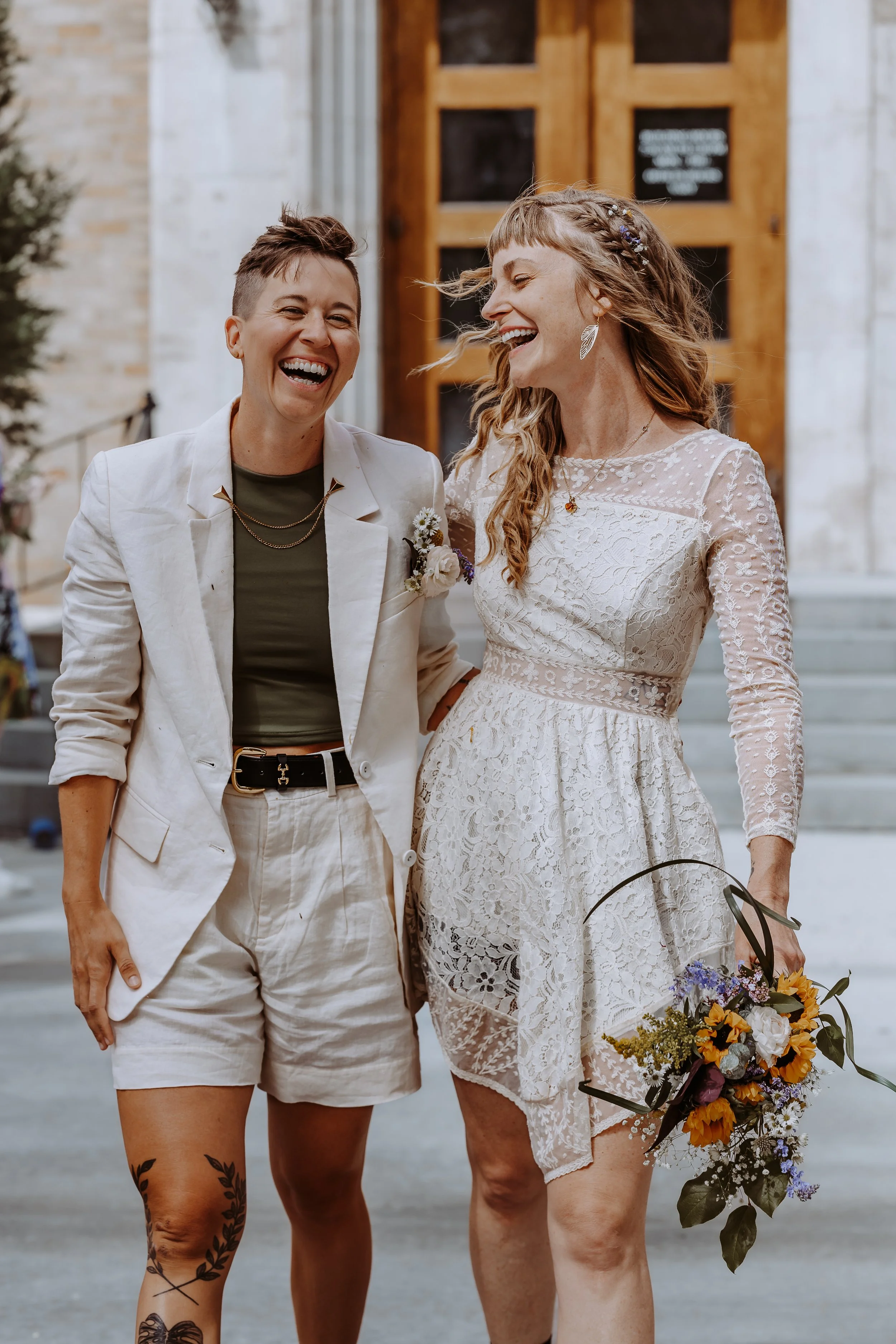 Two women laughing and smiling, standing outdoors in front of a building with wooden accents. One woman has short hair and is wearing a white blazer and matching shorts, with tattoos on her legs. The other woman has long, curly hair, is wearing a white lace dress, and is holding a bouquet of flowers.