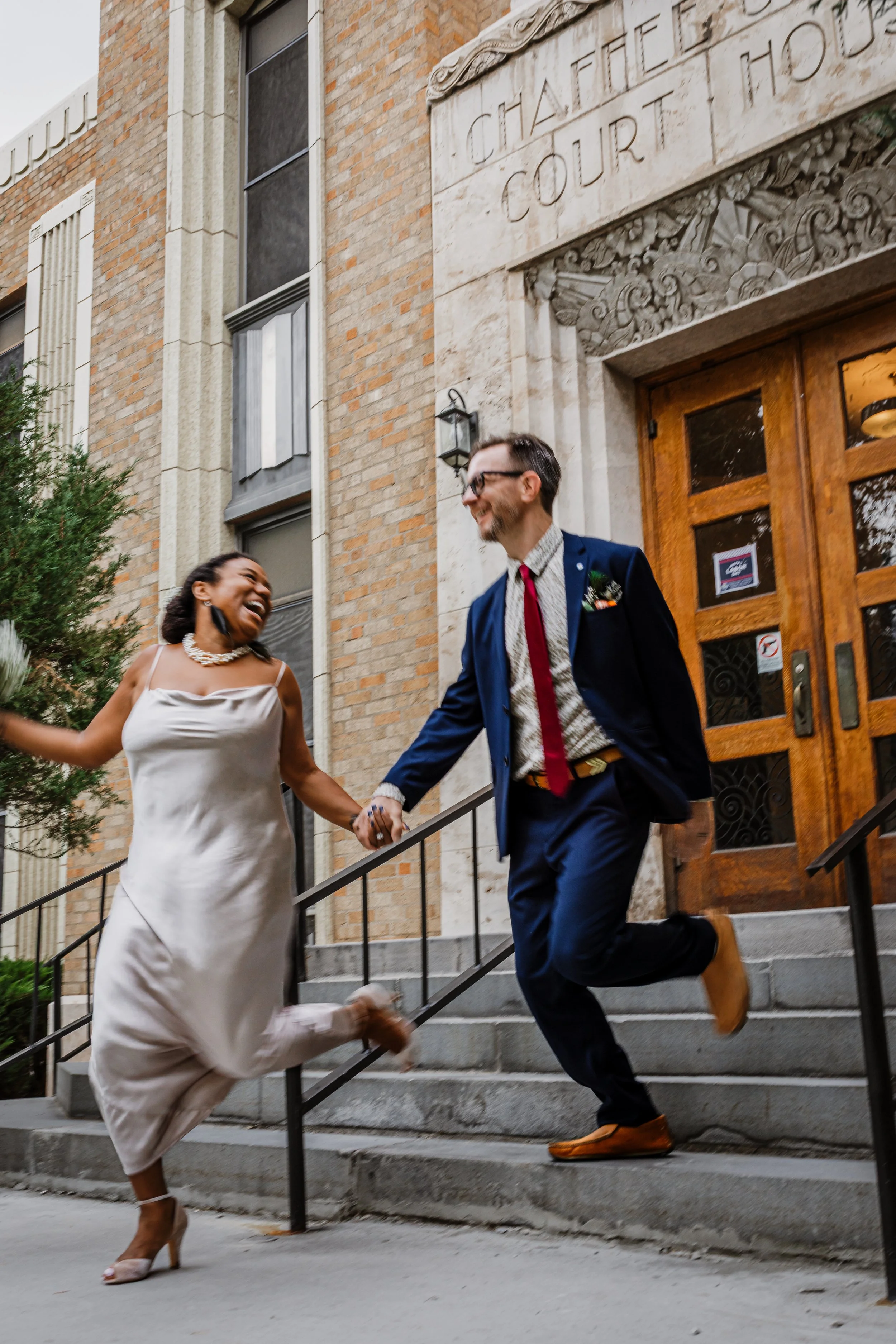 A joyful couple dressed in wedding attire walking down the stairs outside a courthouse, holding hands and smiling.