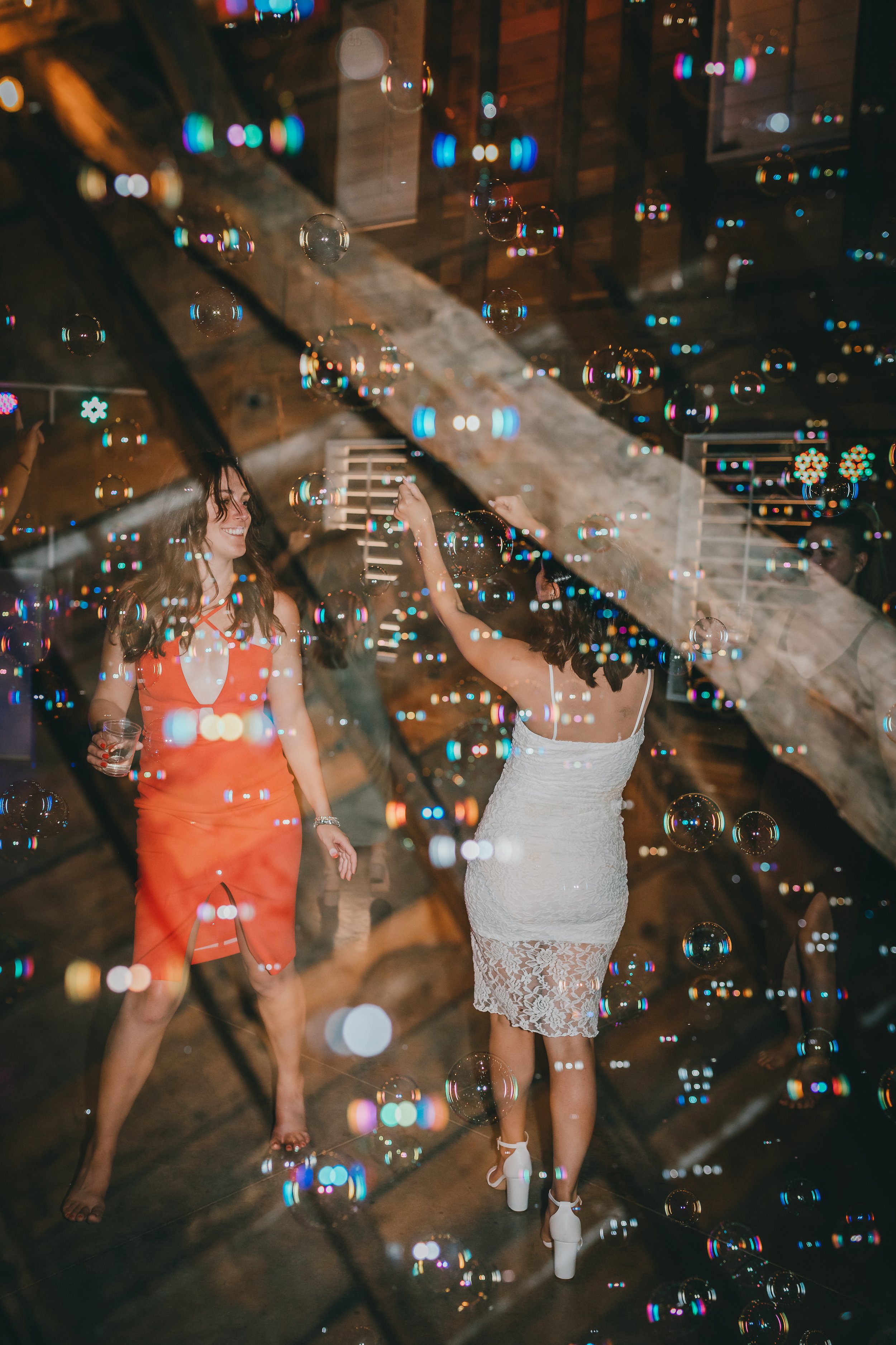 Two women dancing at a celebration party, one in a white lace dress and the other in an orange dress, with bubbles and a wooden interior background.