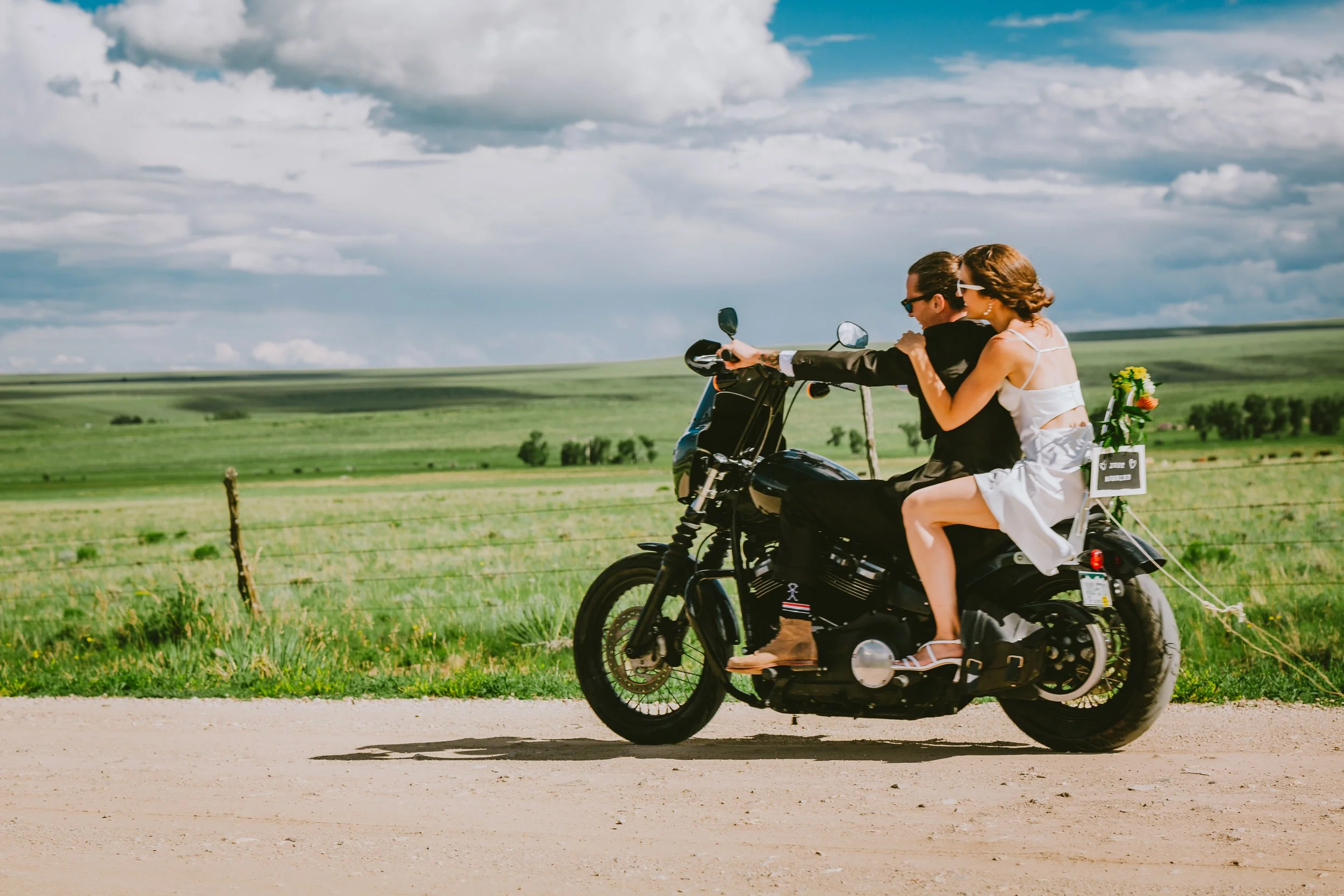 A man and woman riding a motorcycle in a scenic countryside with green fields and a cloudy sky. The woman sitting on back holding a bouquet of flowers and wearing sunglasses.