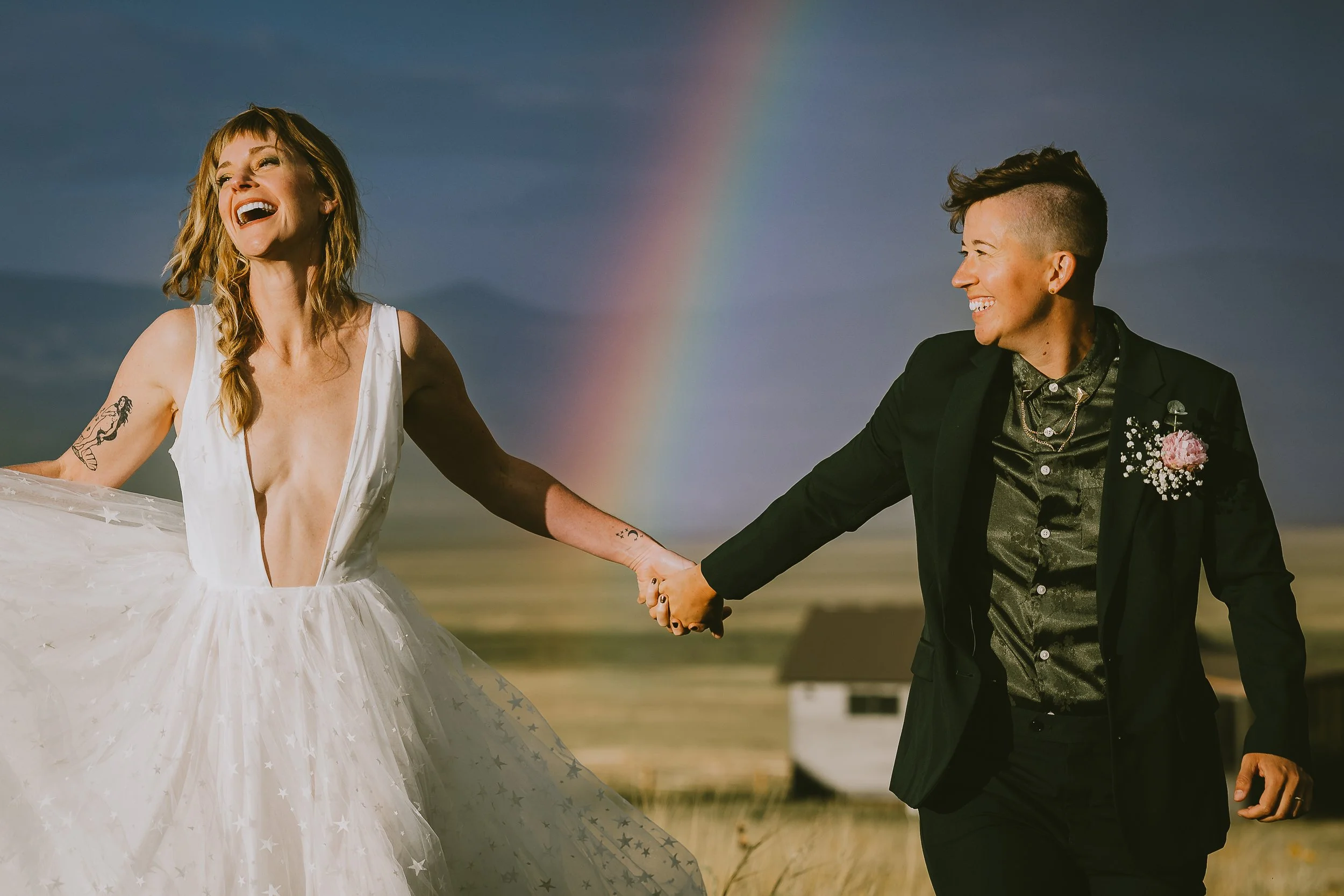 A happy couple holding hands on a beach with a rainbow in the background, the woman wearing a white dress with star patterns and the man in a black suit with a pink flower boutonniere.