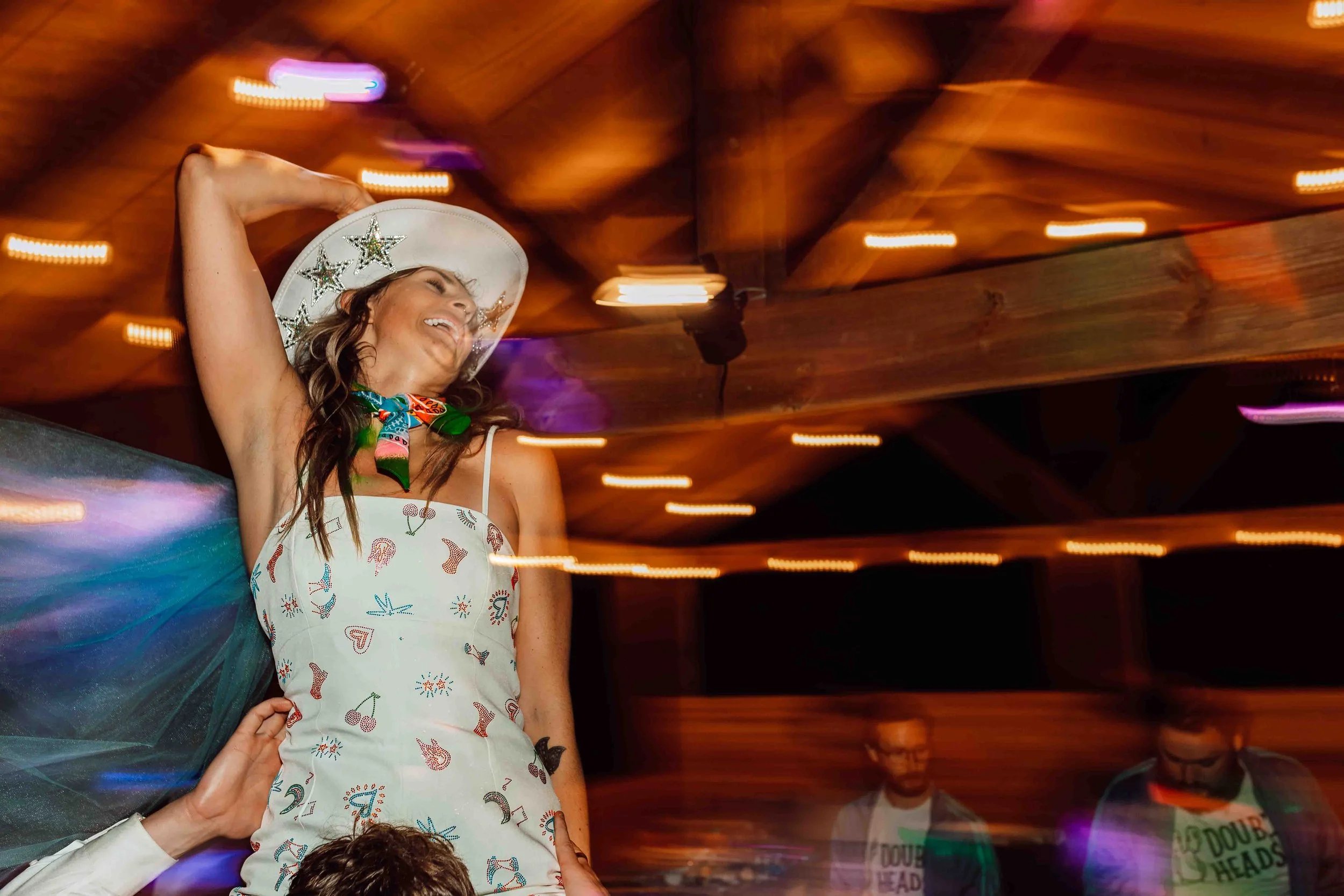 Woman smiling and dressed in a white dress with colorful embroidered designs, wearing a white hat with star decorations, and bright iridescent wings, at a lively indoor event with blurred lights and other people in the background.