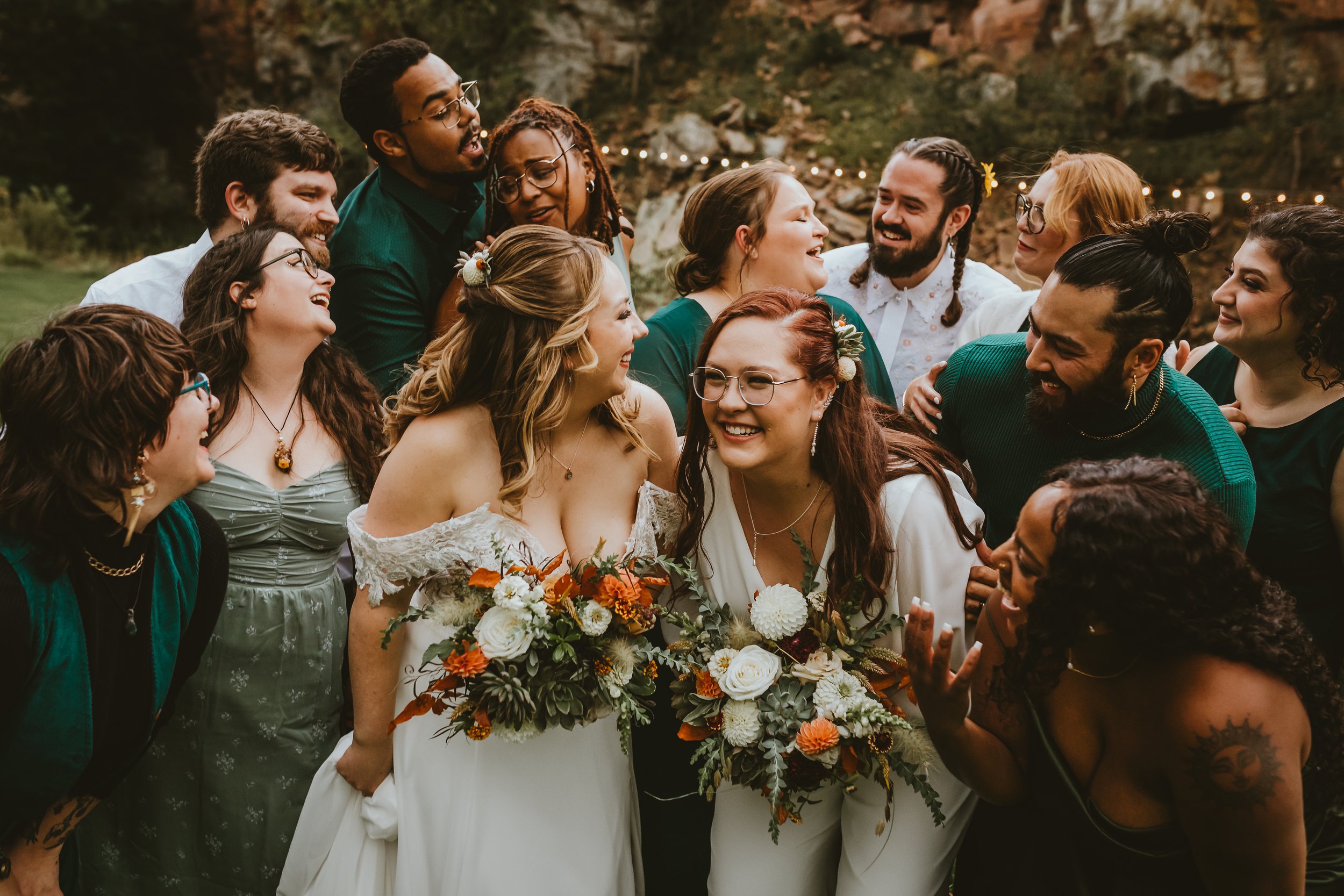 A group of people gathered together outdoors at sunset, smiling and talking, with a wedding couple holding bouquets surrounded by friends.