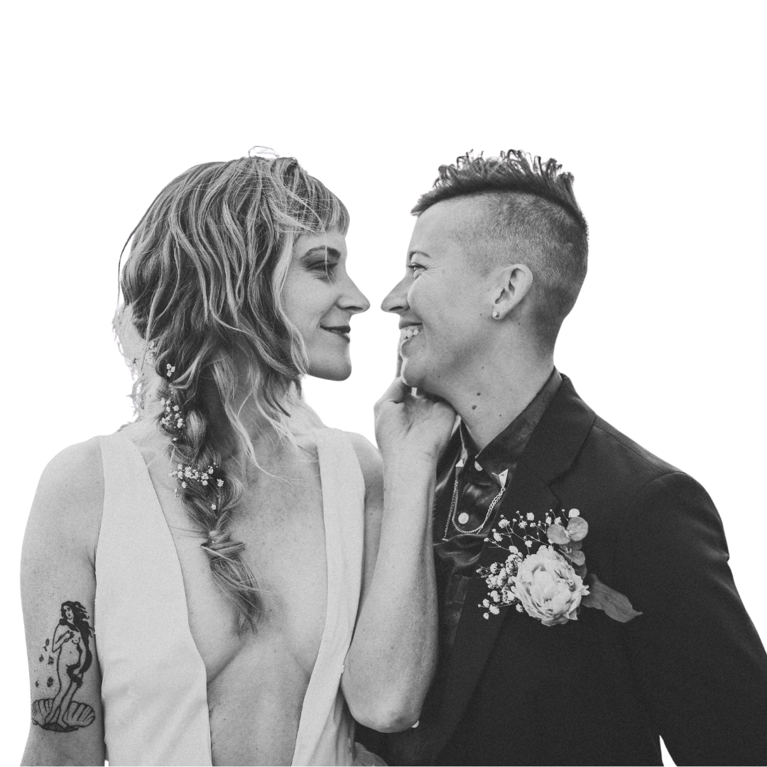 Black and white photo of two women smiling and touching faces, one with curly hair and tattoos, the other with a short haircut, dressed in formal wear, against a black background.