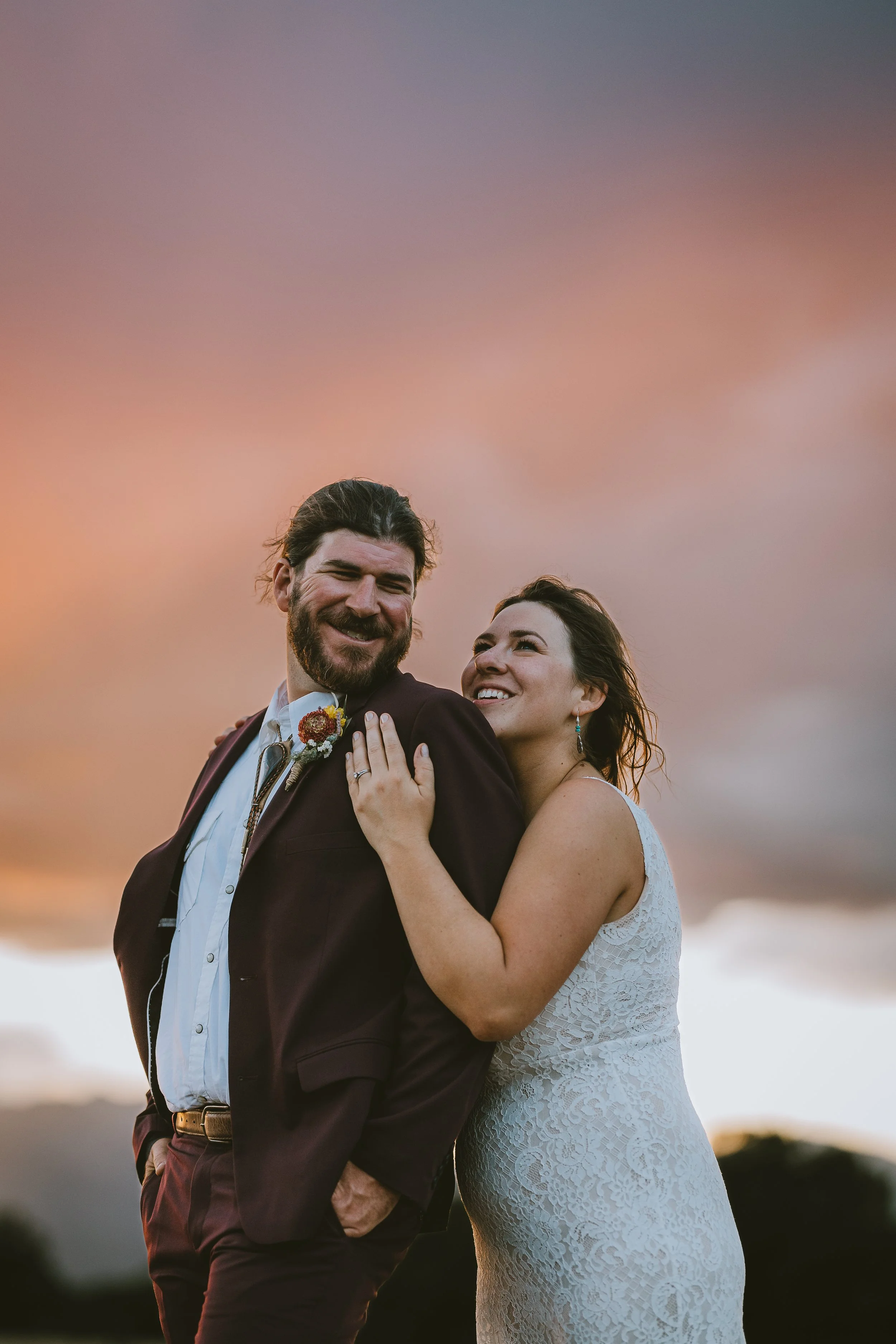 A couple dressed in wedding attire standing close together during sunset, with the woman resting her head on the man's shoulder and both smiling.