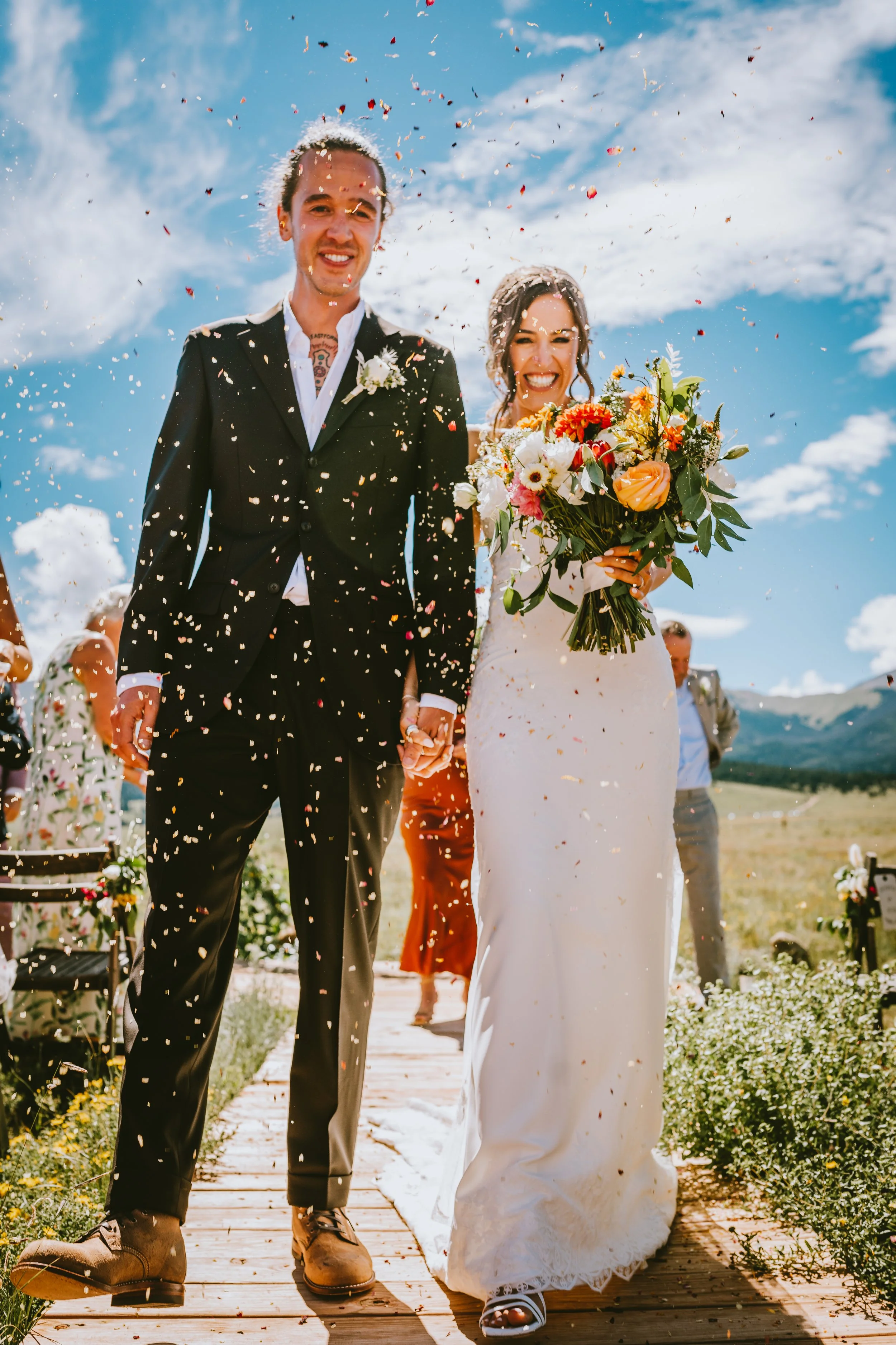A bride and groom holding hands and celebrating their wedding outdoors on a sunny day with bright blue sky and clouds, colorful confetti falling around them, and nature in the background.