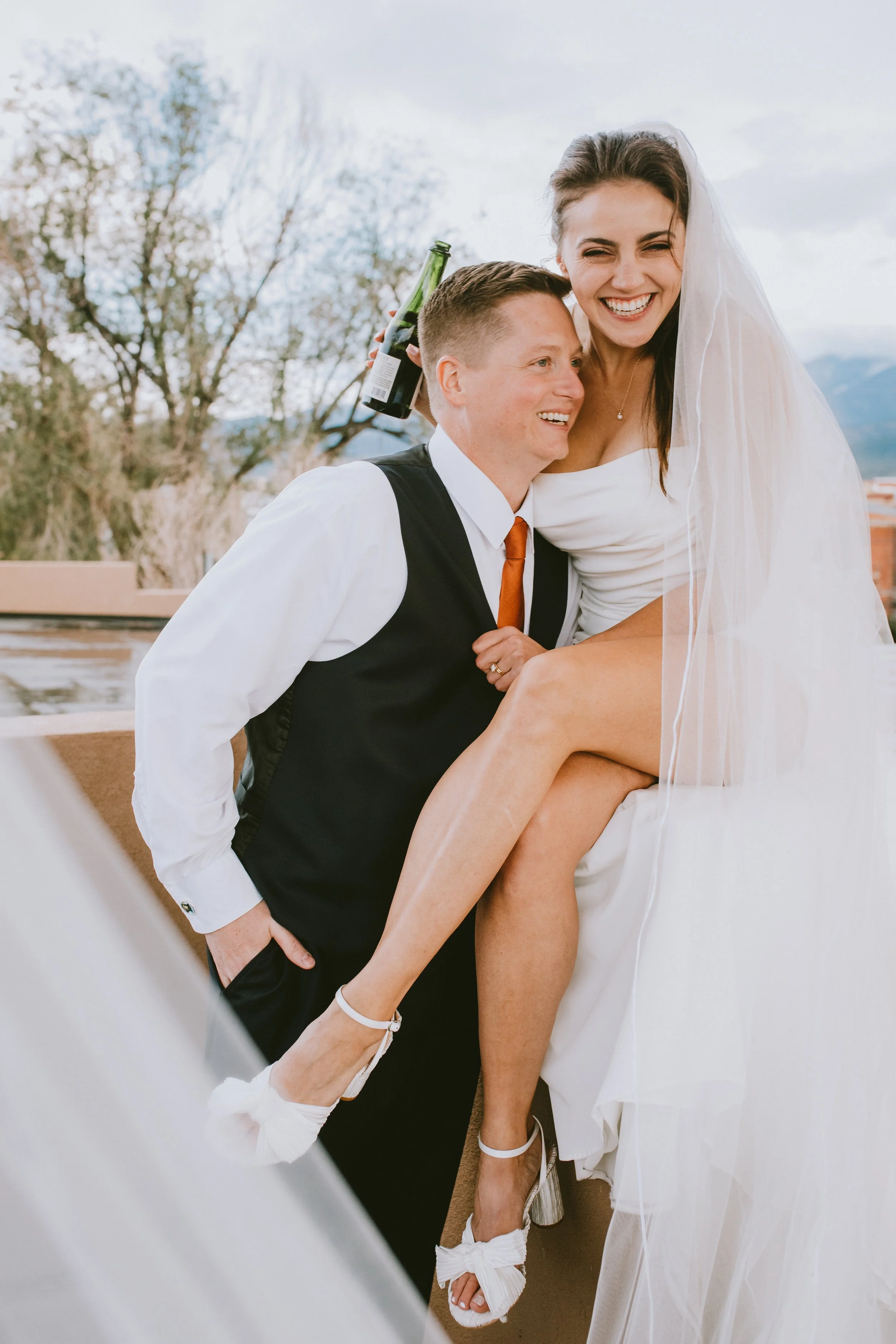 A bride and groom pictured during their wedding celebration. The bride, with dark hair styled up and wearing white, is smiling happily. The groom, with short hair, is also smiling and dressed in a white shirt, black vest, and tie. The groom is holding a bottle of champagne while the bride is sitting on his lap with one leg over his shoulder, showing her white wedding dress and high-heeled shoes.