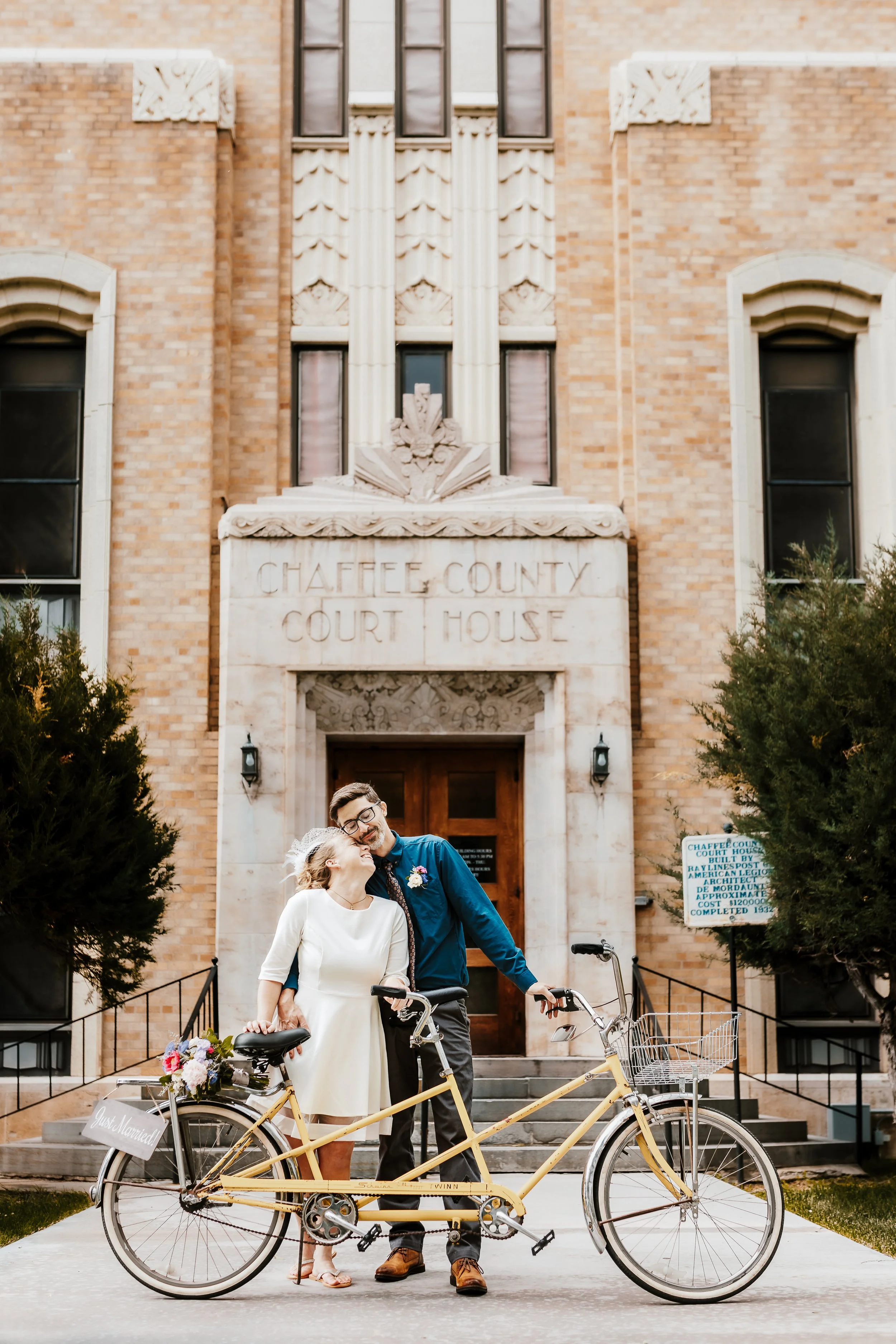 A newlywed couple with bicycles standing in front of the Chaffee County Courthouse, smiling and sharing a moment together.