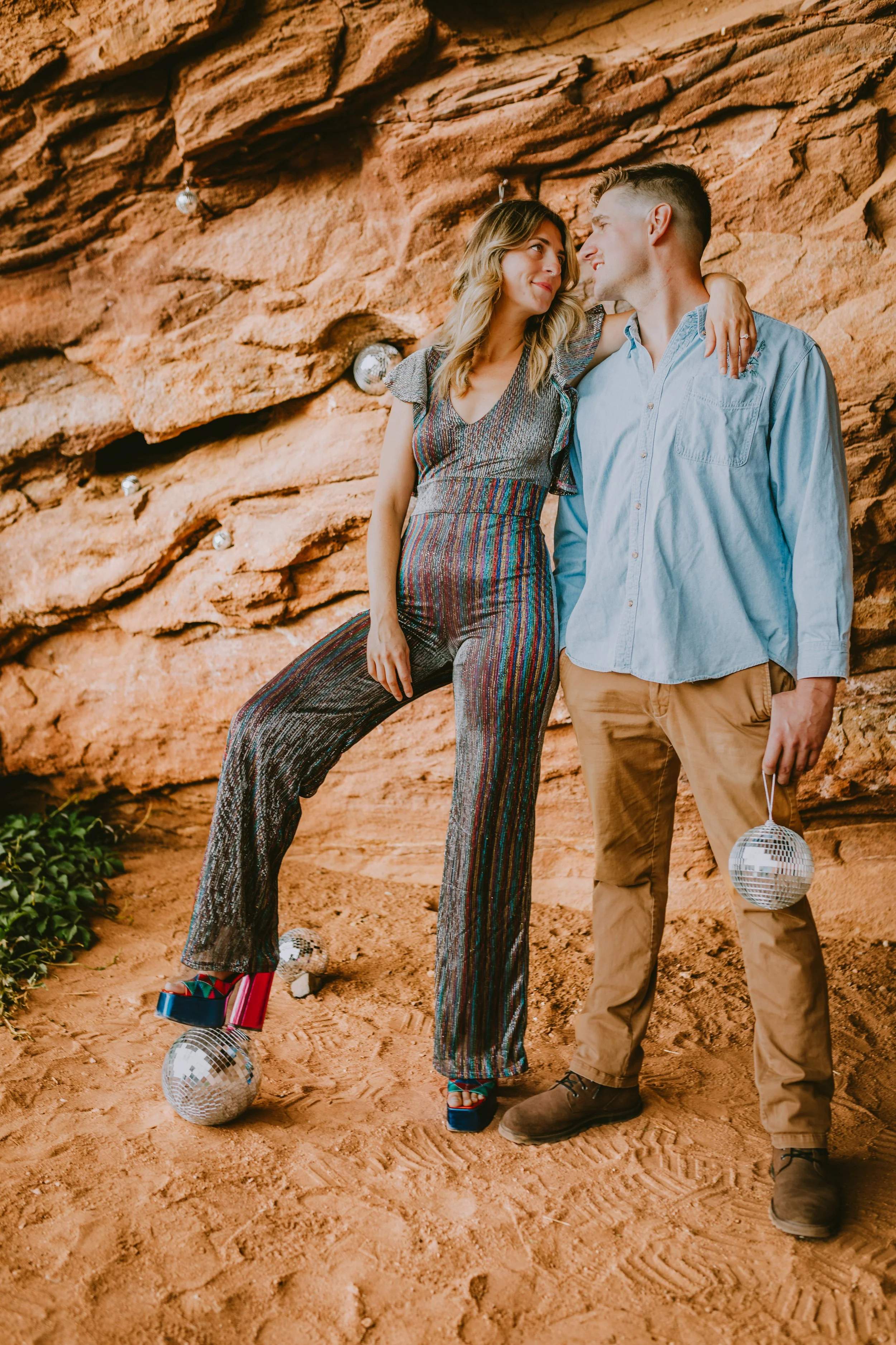 A couple standing on a dusty ground with disco balls and crates, looking into each other's eyes, near a rock wall backdrop.