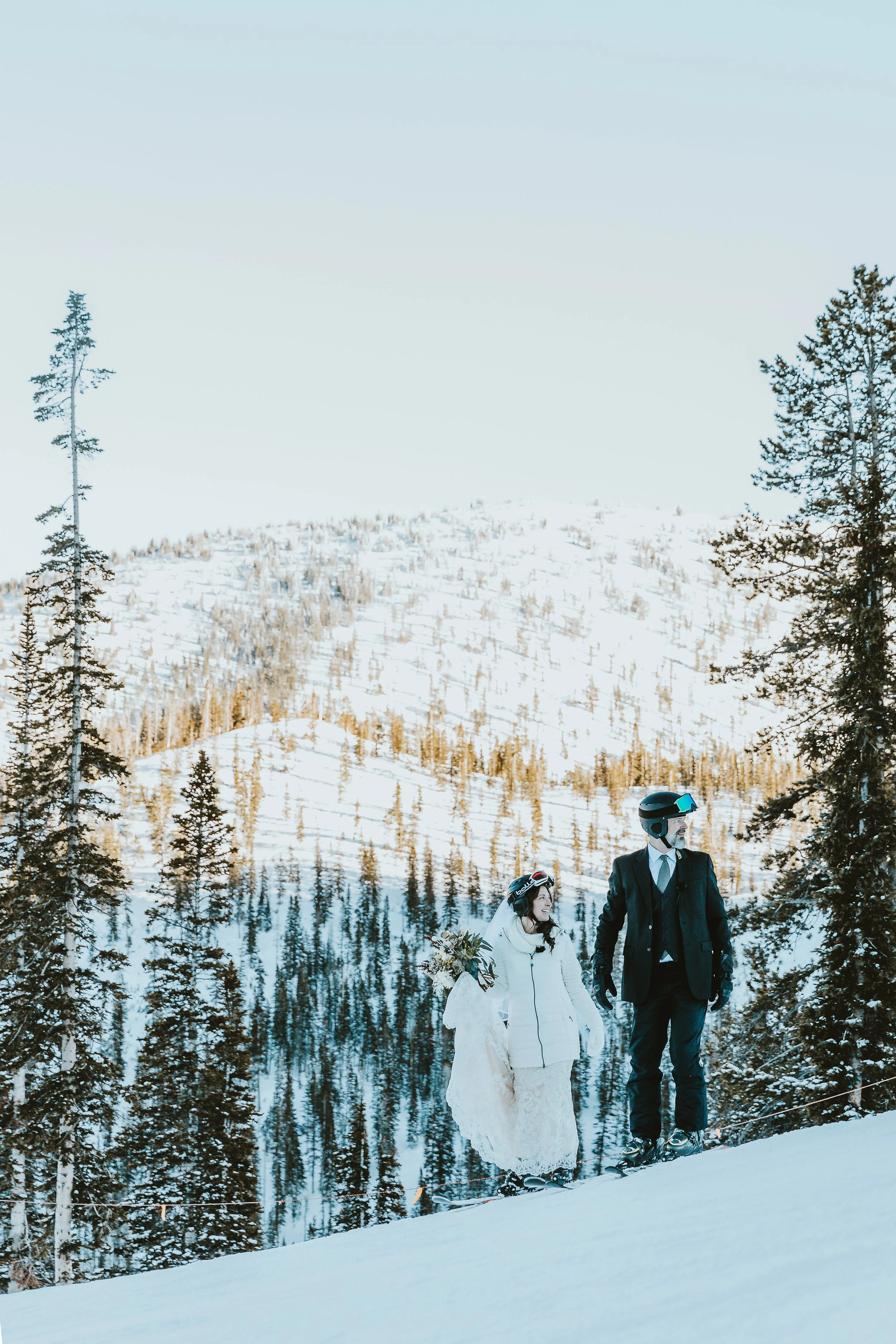 Couple holding hands on snowy mountain with skis on. Bride holding dress and bouquet in one hand.  Ski wedding, Salida, CO.