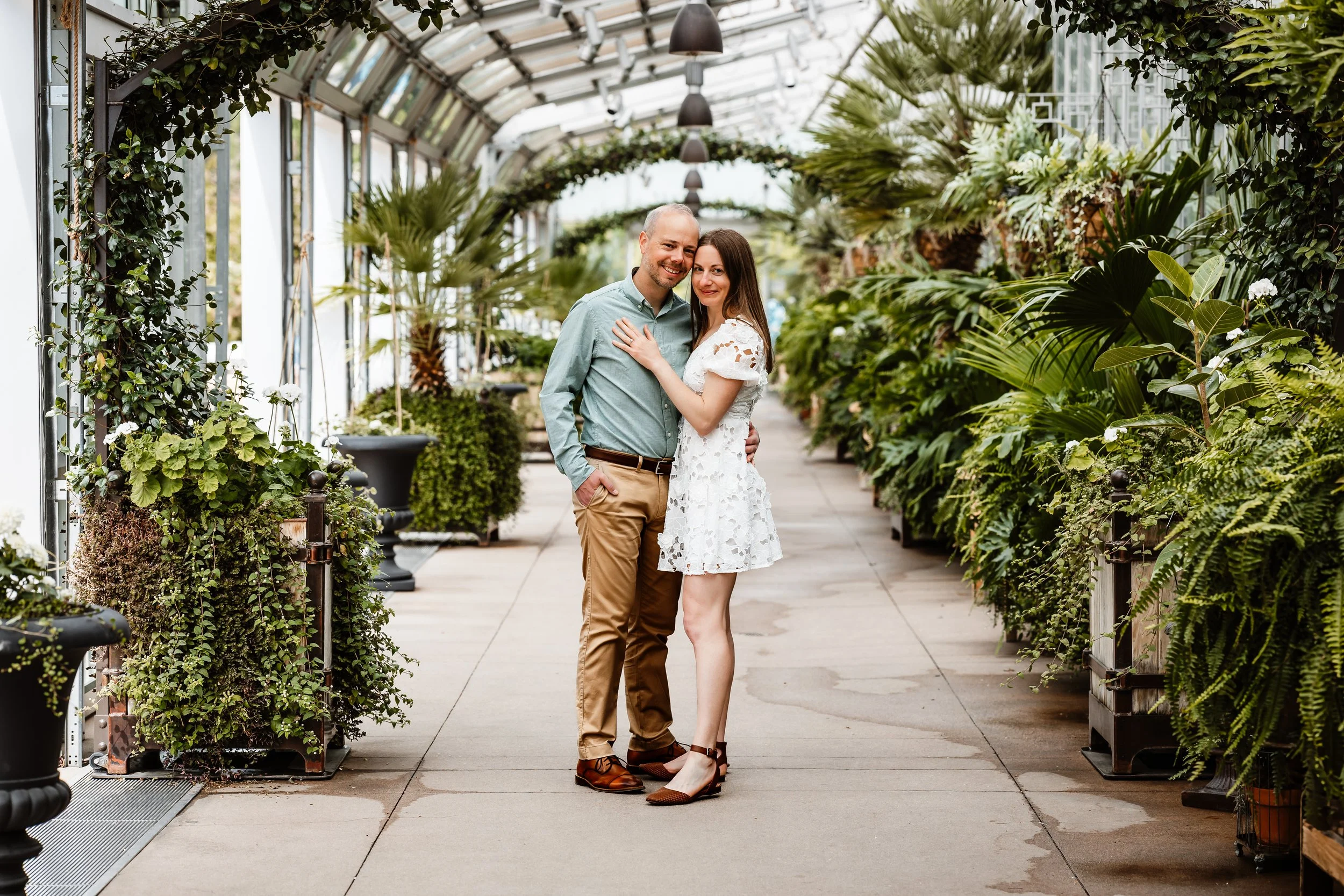 Couple during their engagement session at the Denver Botanical Gardens. Holding each other close smiling at the camera surrounded by greenery in an arch way.