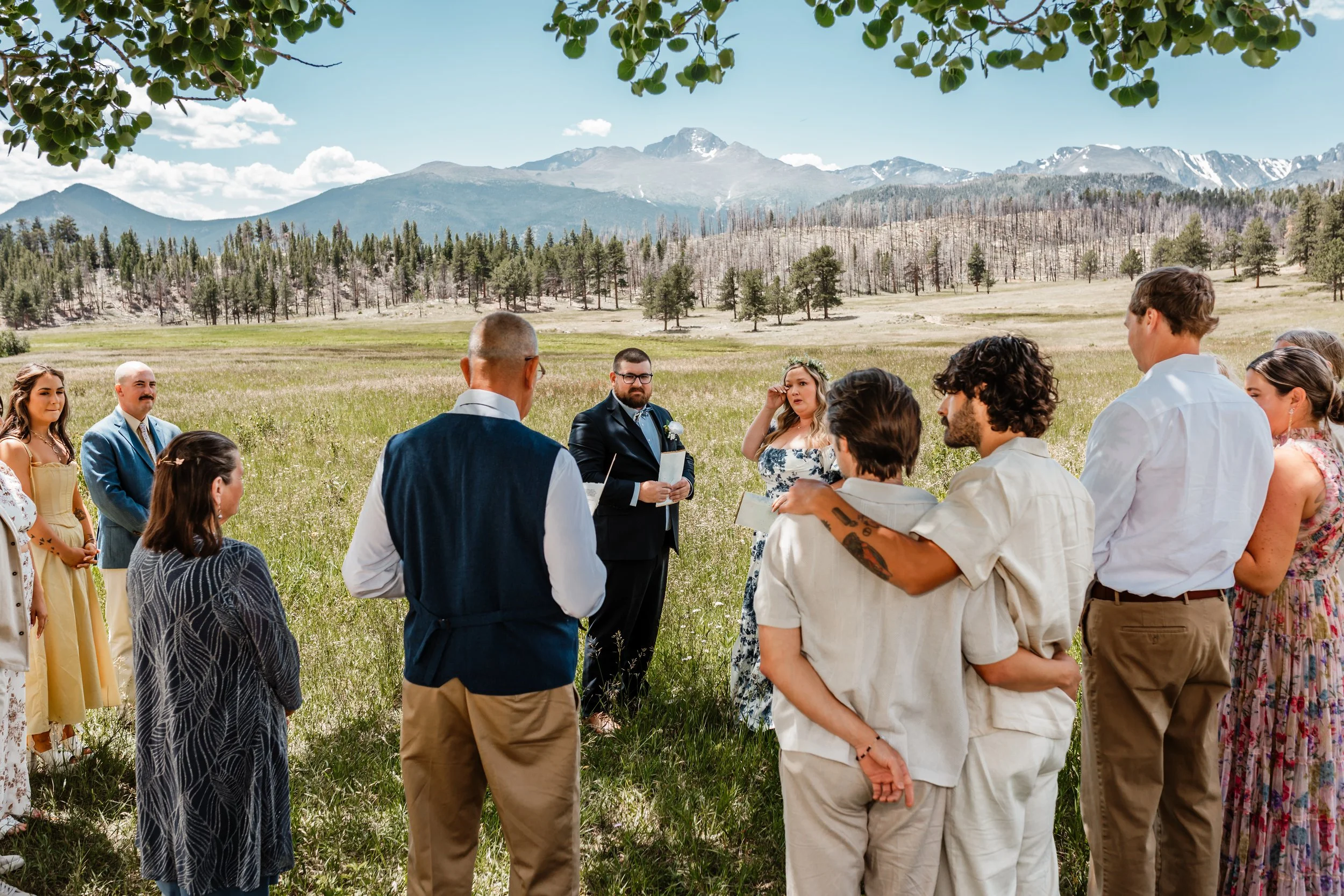 Couple standing with their small ceremony guests at Rocky Mountain National Park under a shade tree. Mountains in the background, the wedding guests are all standing with their backs turned to the photographer forming a semi-circle around the couple.