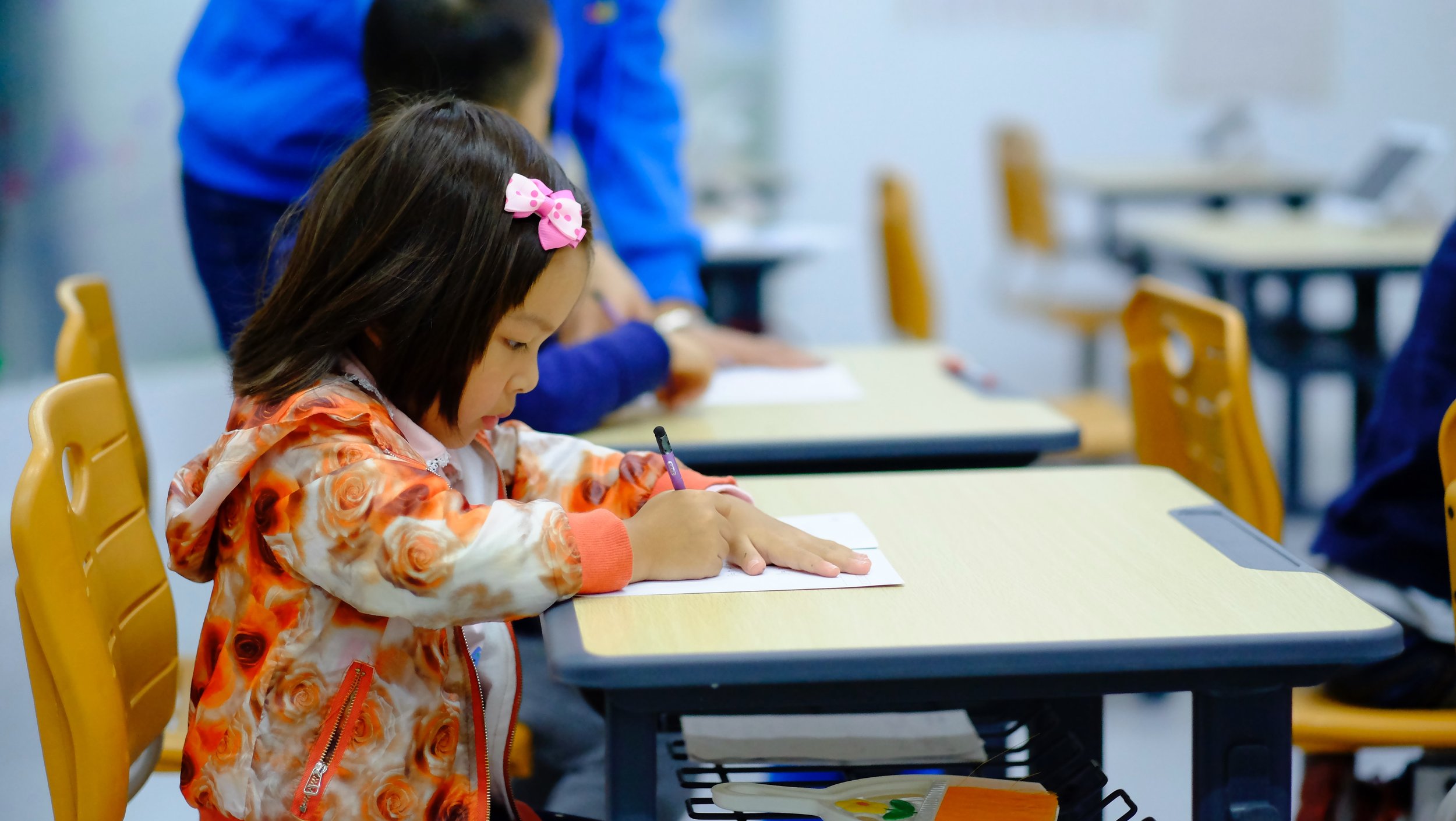Child writing at a school desk with a pencil, wearing a floral jacket and a pink hair bow, in a classroom setting.