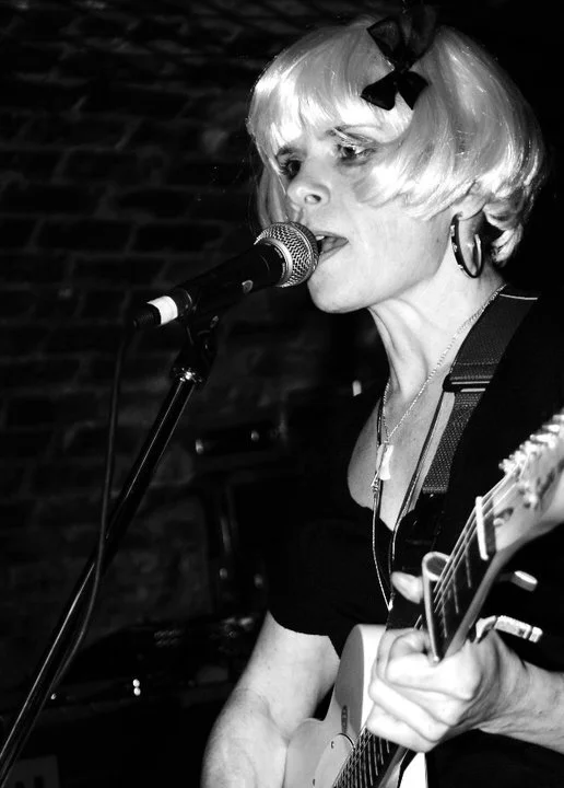 A woman with short blonde hair, wearing a black bow hair accessory and large hoop earrings, sings into a microphone while playing an acoustic guitar in a dimly lit setting.