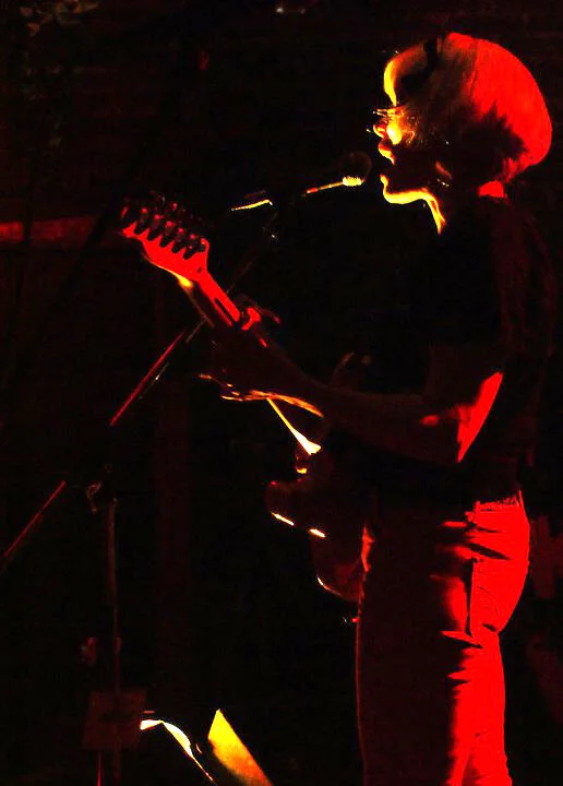 A woman singing and playing an electric guitar on stage with dramatic red and yellow lighting.