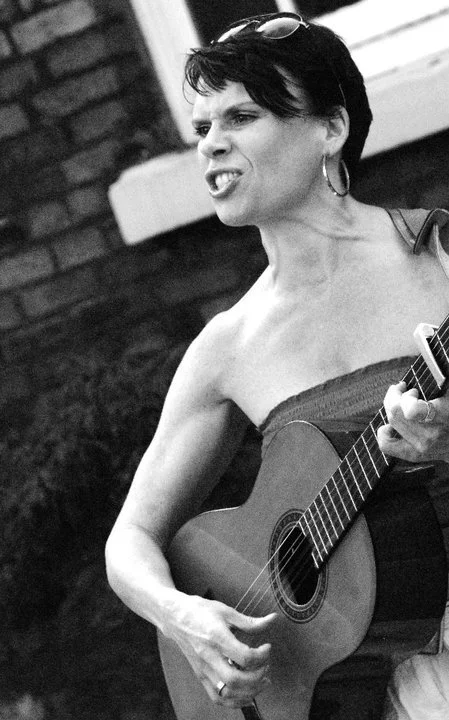 A woman with short dark hair, wearing glasses on her head, earrings, and a strapless top, playing an acoustic guitar outdoors.