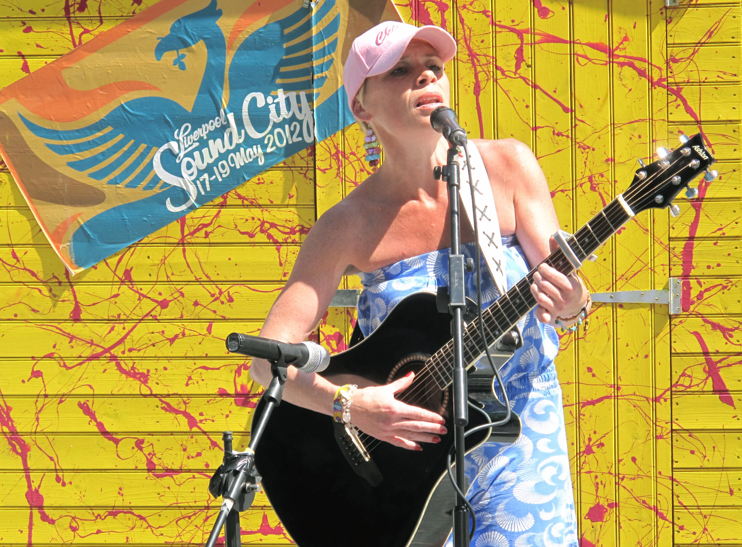 A woman wearing a pink baseball cap, earrings, and a blue and white patterned sleeveless top is singing into a microphone while playing an acoustic guitar. Behind her, there is a colorful yellow background with pink splatters and a blue and white ban