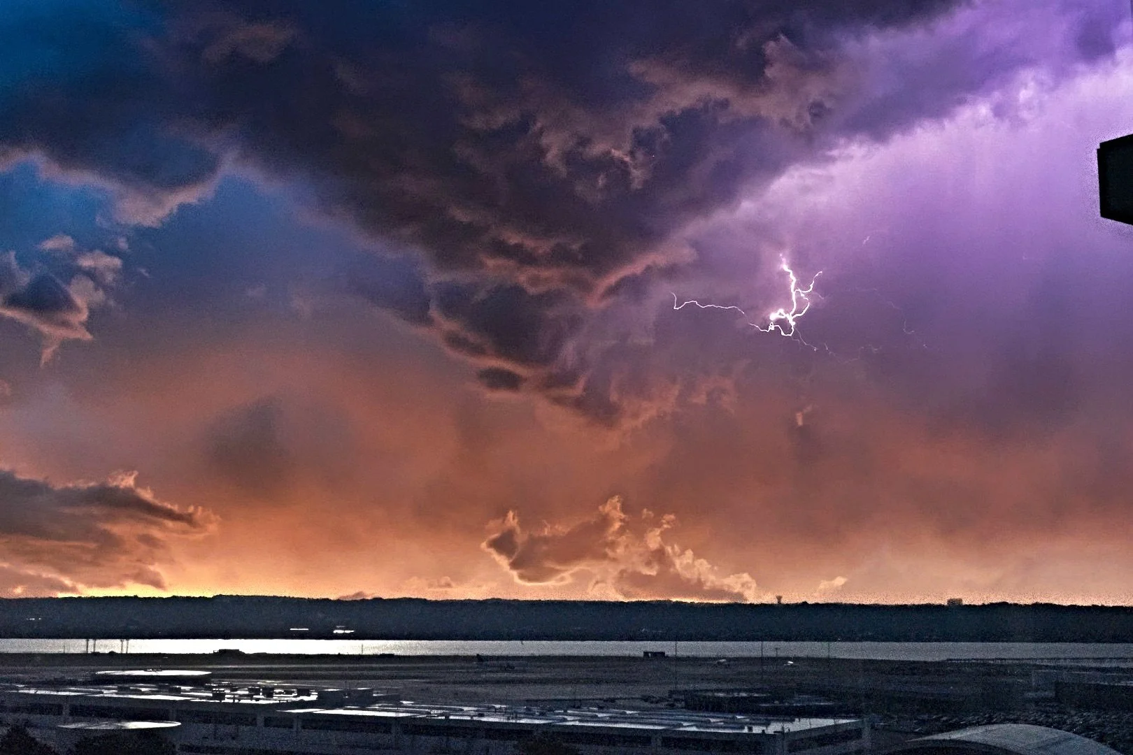 Dramatic sunset with dark storm clouds and a lightning bolt over a silhouette of buildings and water.