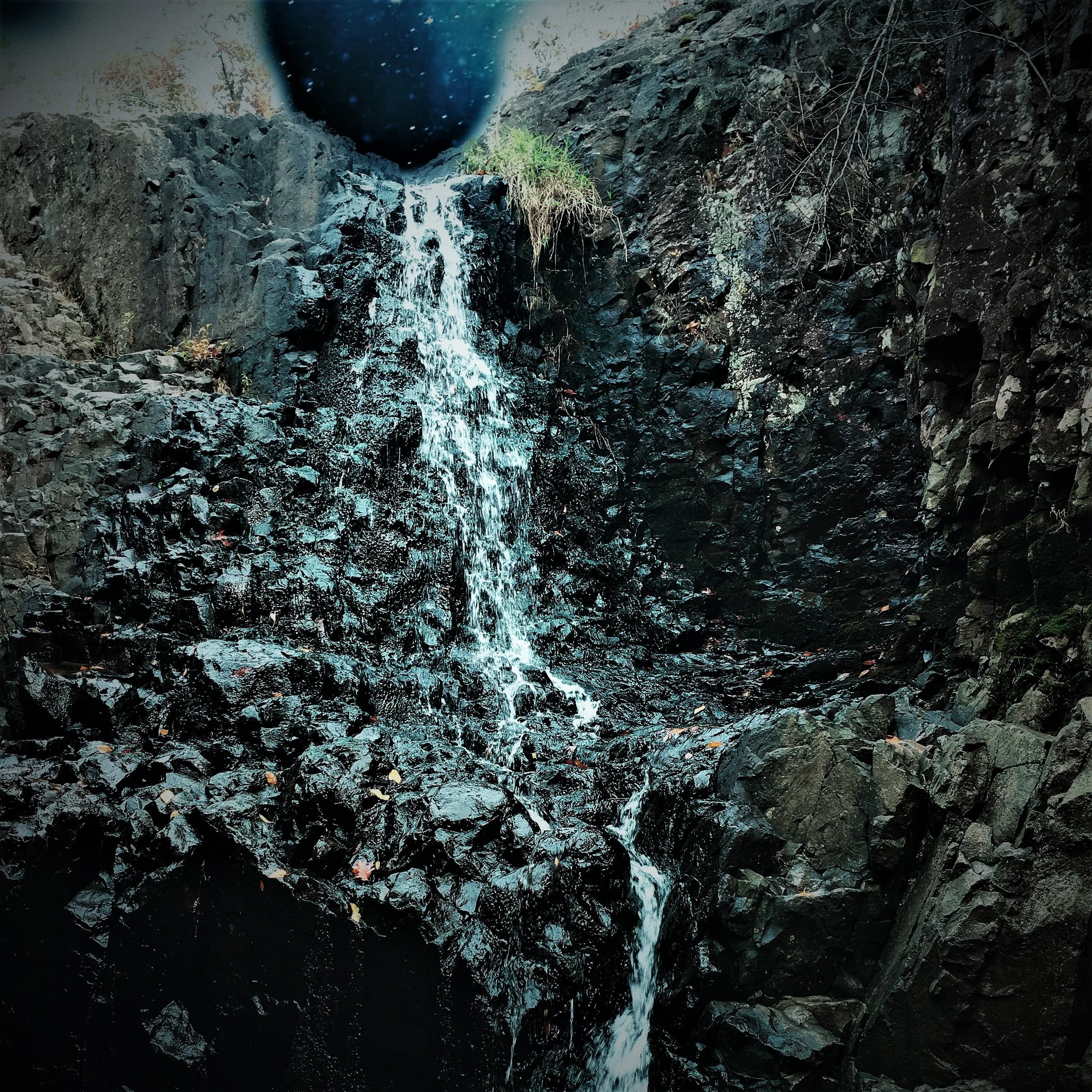 Small waterfall cascading over dark, rocky cliffs with greenery at the top and dark sky above.