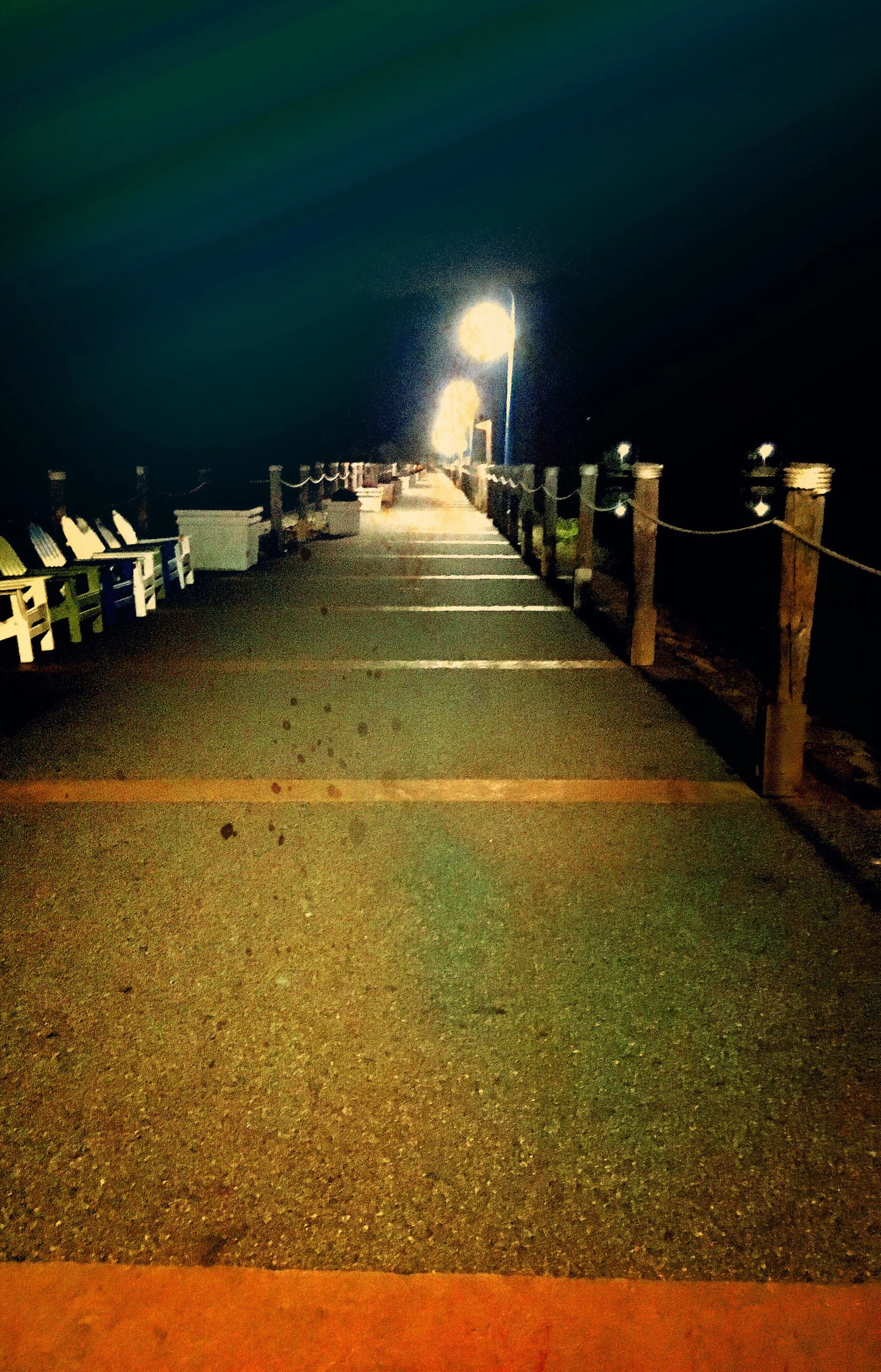 Nighttime view of a long pier with lampposts illuminating the path, lined with benches and bordered by rope railings.