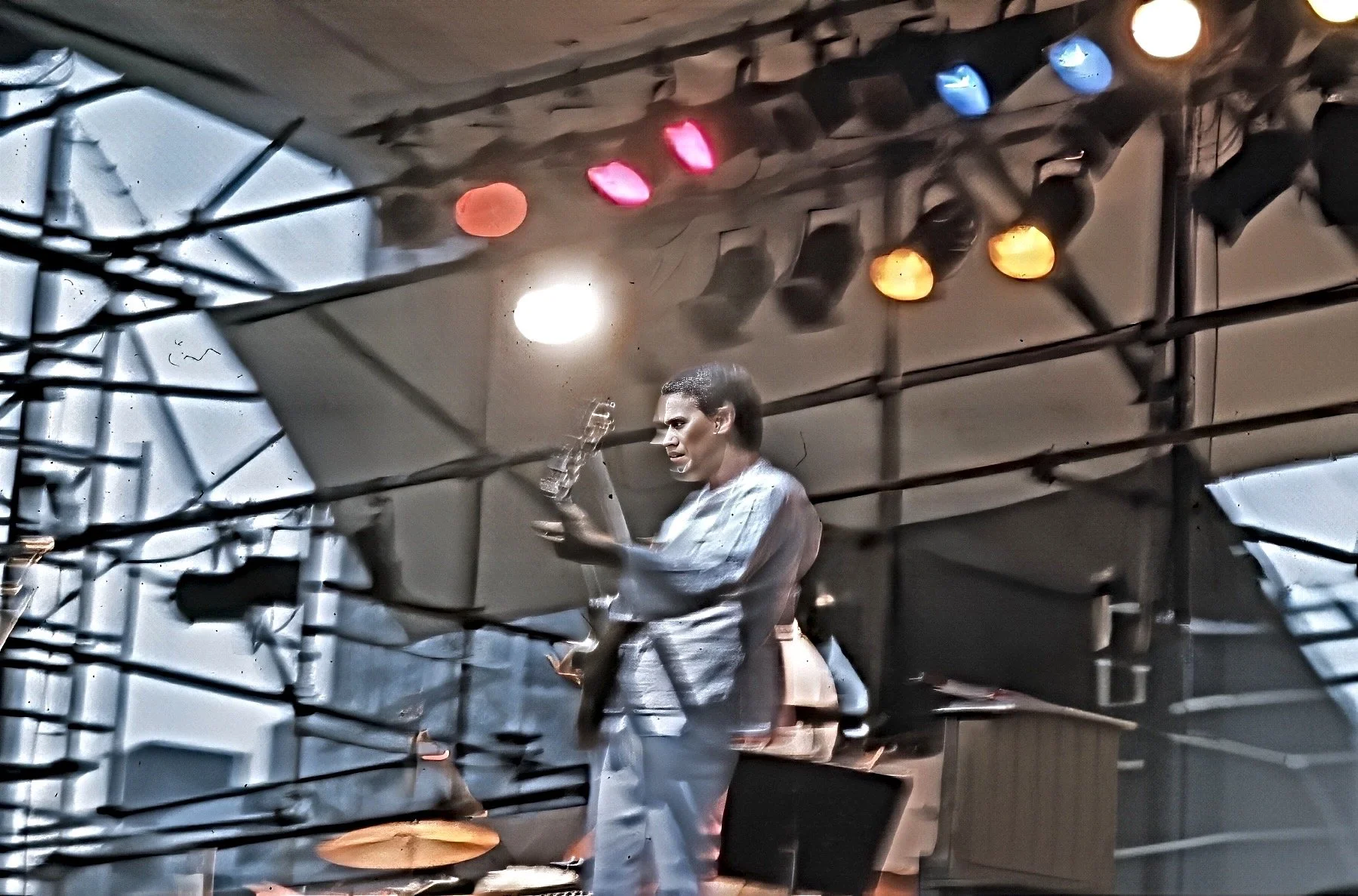 A person with dark hair playing an electric guitar on a stage with colorful stage lights overhead, reflected in a mirror.