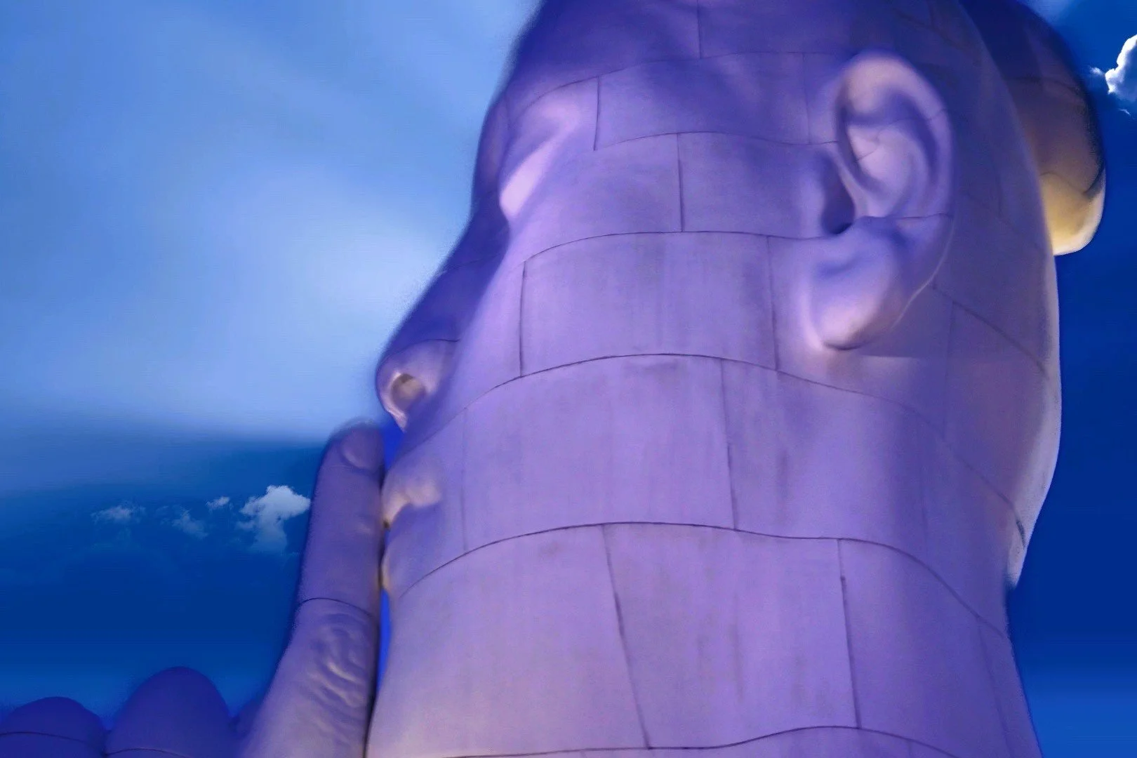 Stone sculpture of a face with a finger on the lips, resembling a gesture for silence, against a blue sky.