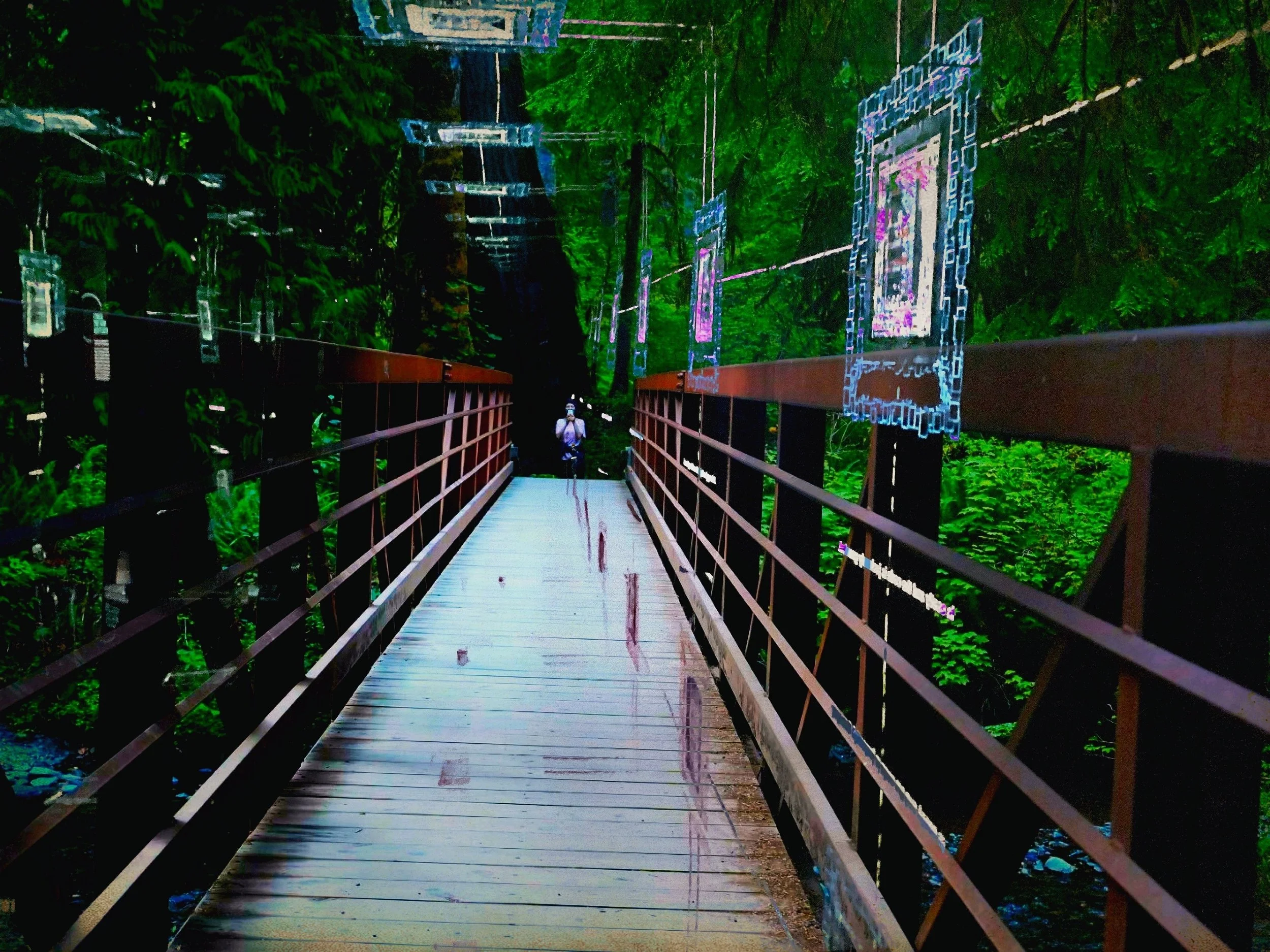 A wooden pedestrian bridge with metal railings in a forest setting. Art installations or frames hang above the bridge, creating an artistic atmosphere. Lush greenery surrounds the scene. A person is visible at the far end of the bridge.