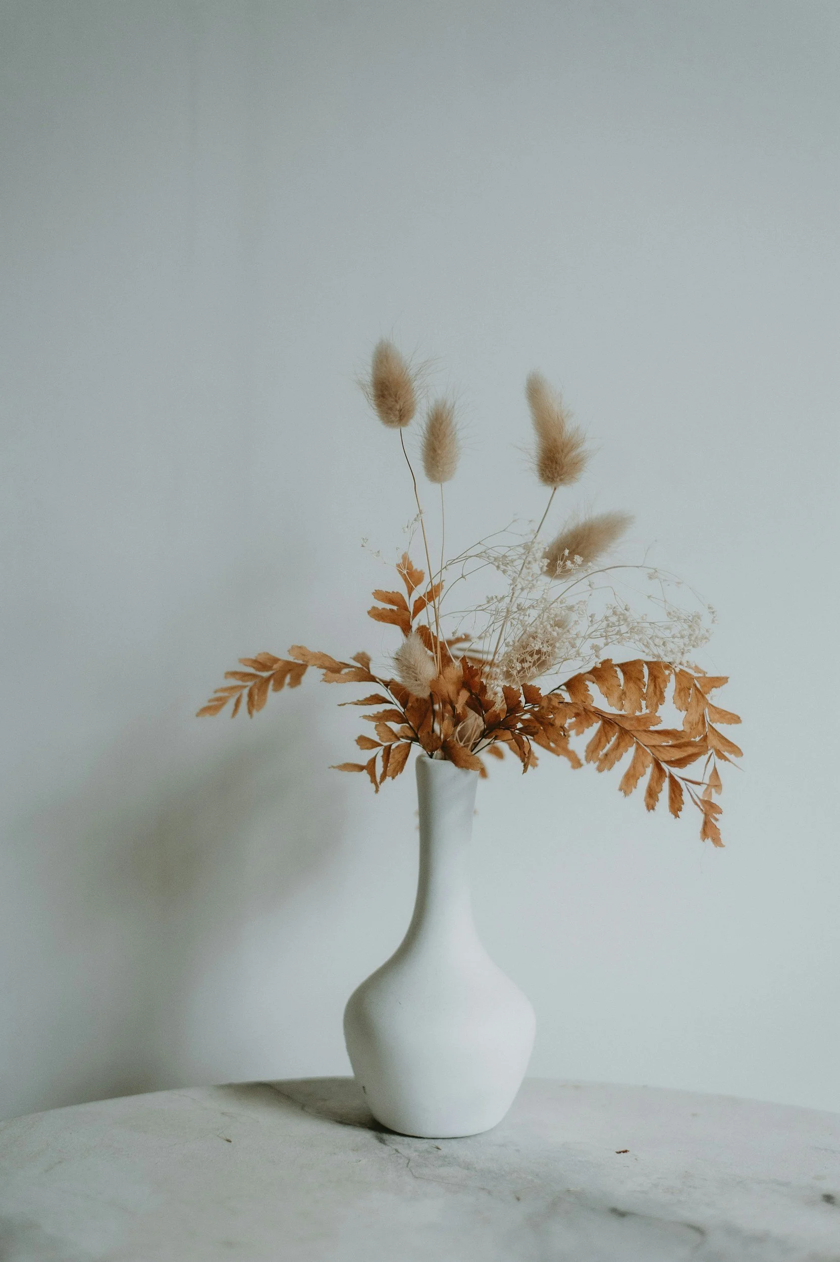 A white vase with an abstract shape holds dried brown leaves and beige fluffy grass stems, placed on a light-colored surface against a plain white wall.