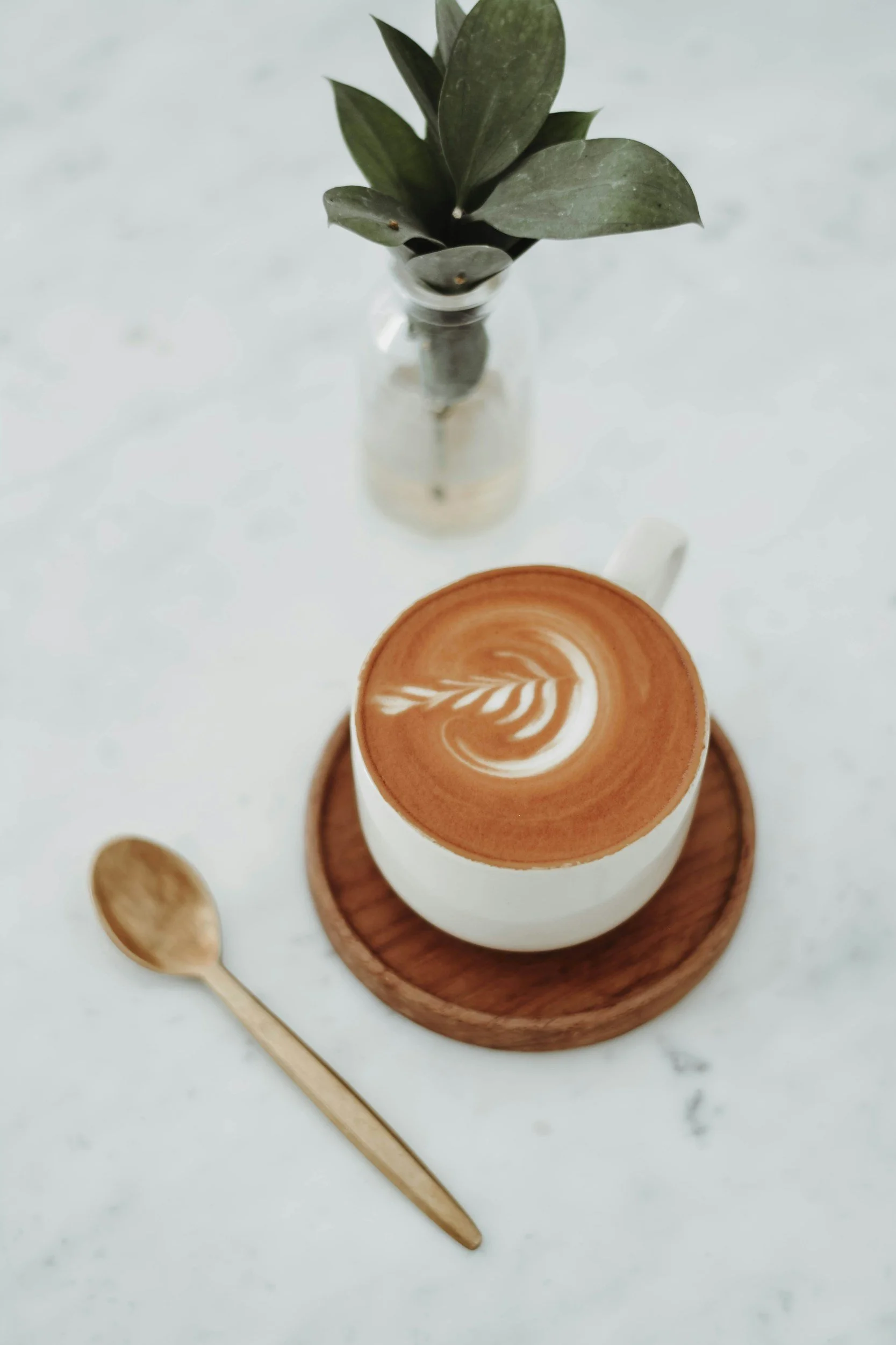 A white coffee mug with a latte art leaf design, placed on a round wooden tray, a small gold spoon beside it, and a small glass vase with green leaves behind it, all on a white marble surface.