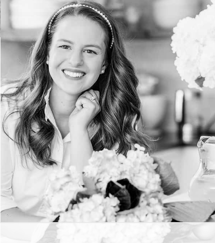 Interior designer and home decor influencer Nina Hendrick with Nina Hendrick Home, smiling, wearing a headband and a white shirt, sitting at a table with a bouquet of flowers and a glass pitcher.