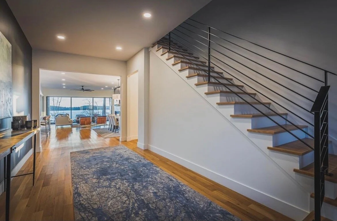 Interior view of a modern home with an open floor plan, hardwood floors, a staircase with wooden steps and metal railing, and a living area with large windows overlooking a lake.