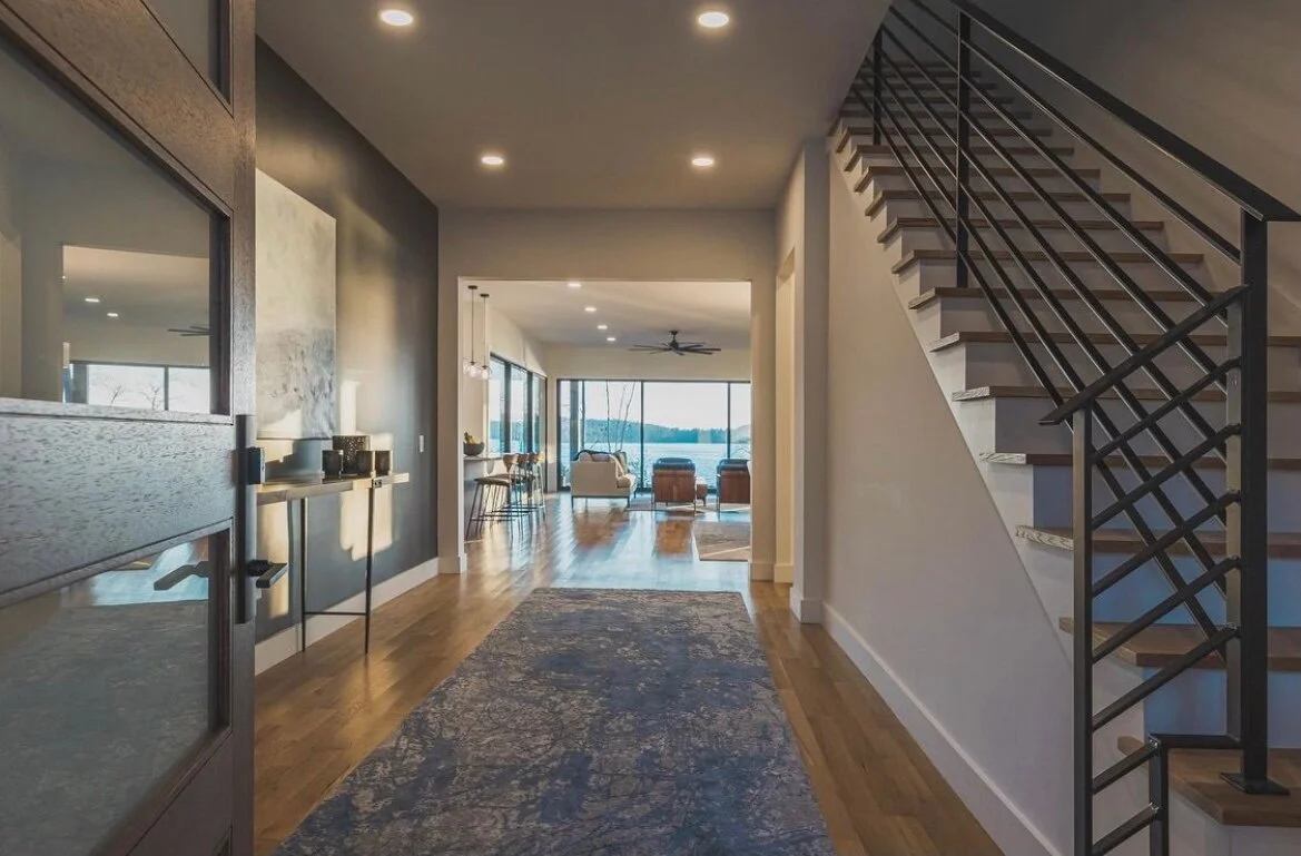 Interior view of a modern home with hardwood floors, a staircase on the right, and a living room with large windows overlooking a body of water in the background.
